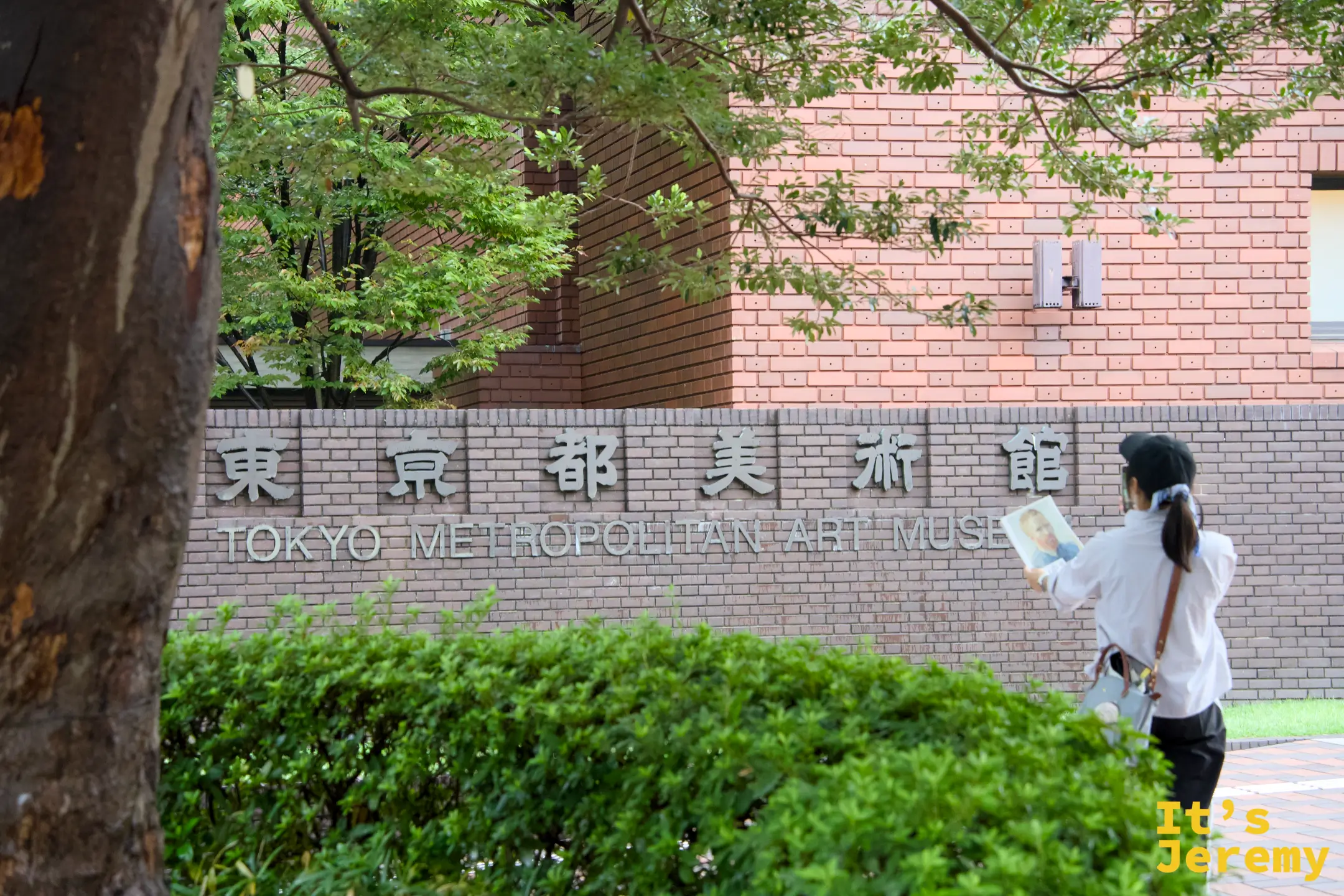 一位女士手持手冊站在東京都美術館外的標示牌旁,標示牌上有「東京都美術館 TOKYO METROPOLITAN ART MUSEUM」的中英文字樣,周圍是茂密的綠樹