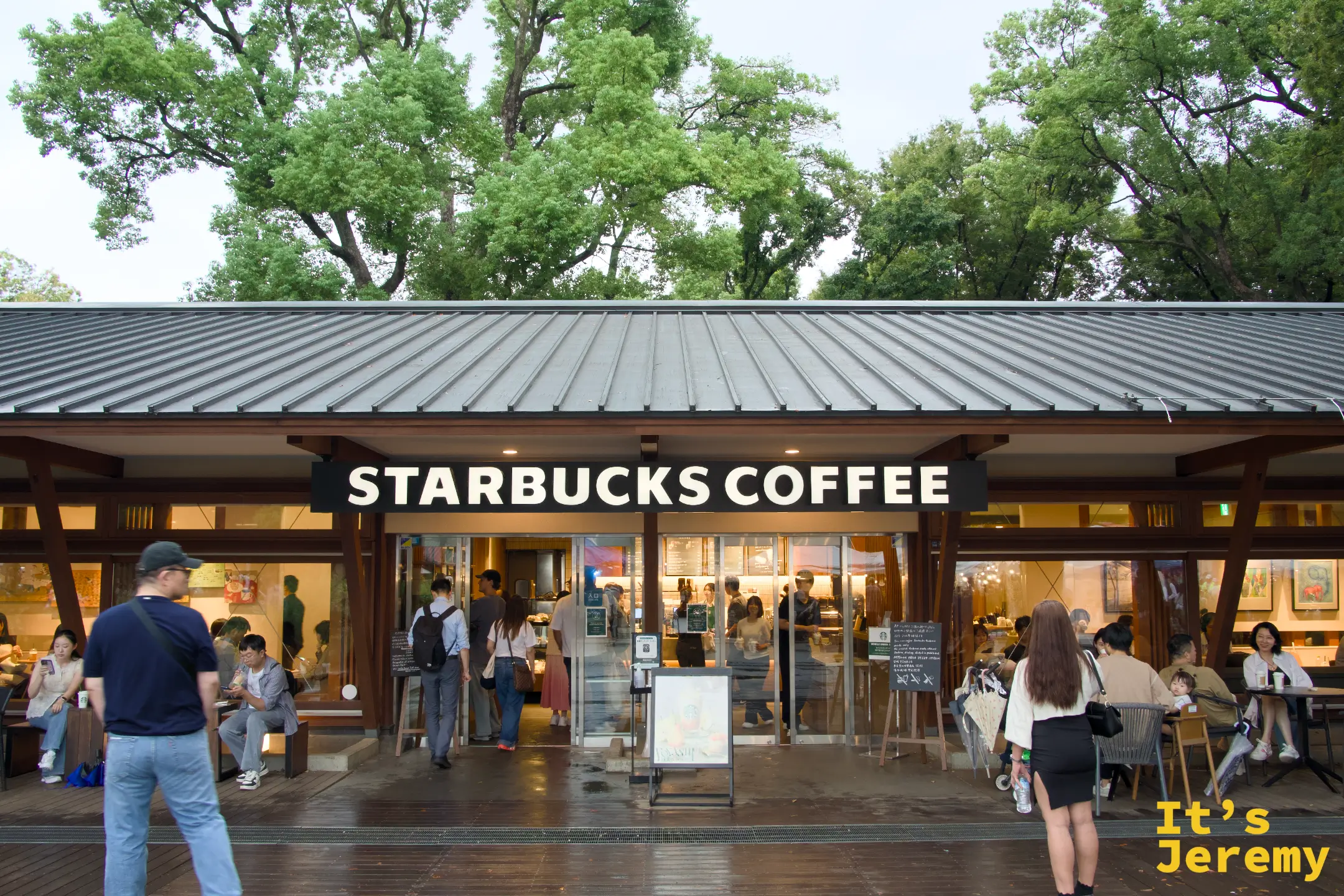 上野公園的星巴克全景,低矮木屋頂建築,「STARBUCKS COFFEE」招牌清晰,戶外座位坐著客人,背後是公園高大的老樹