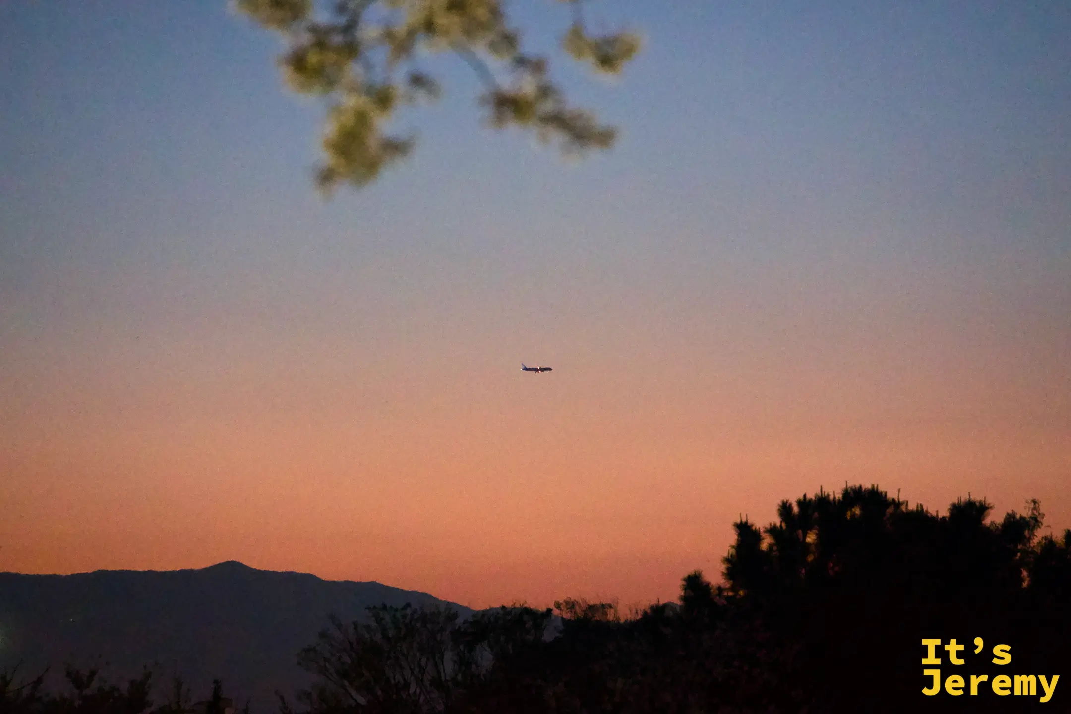 夕陽餘霞染紅整片天空，遠處的山稜線壓低成剪影，一架飛機悄悄穿越橘色的天幕