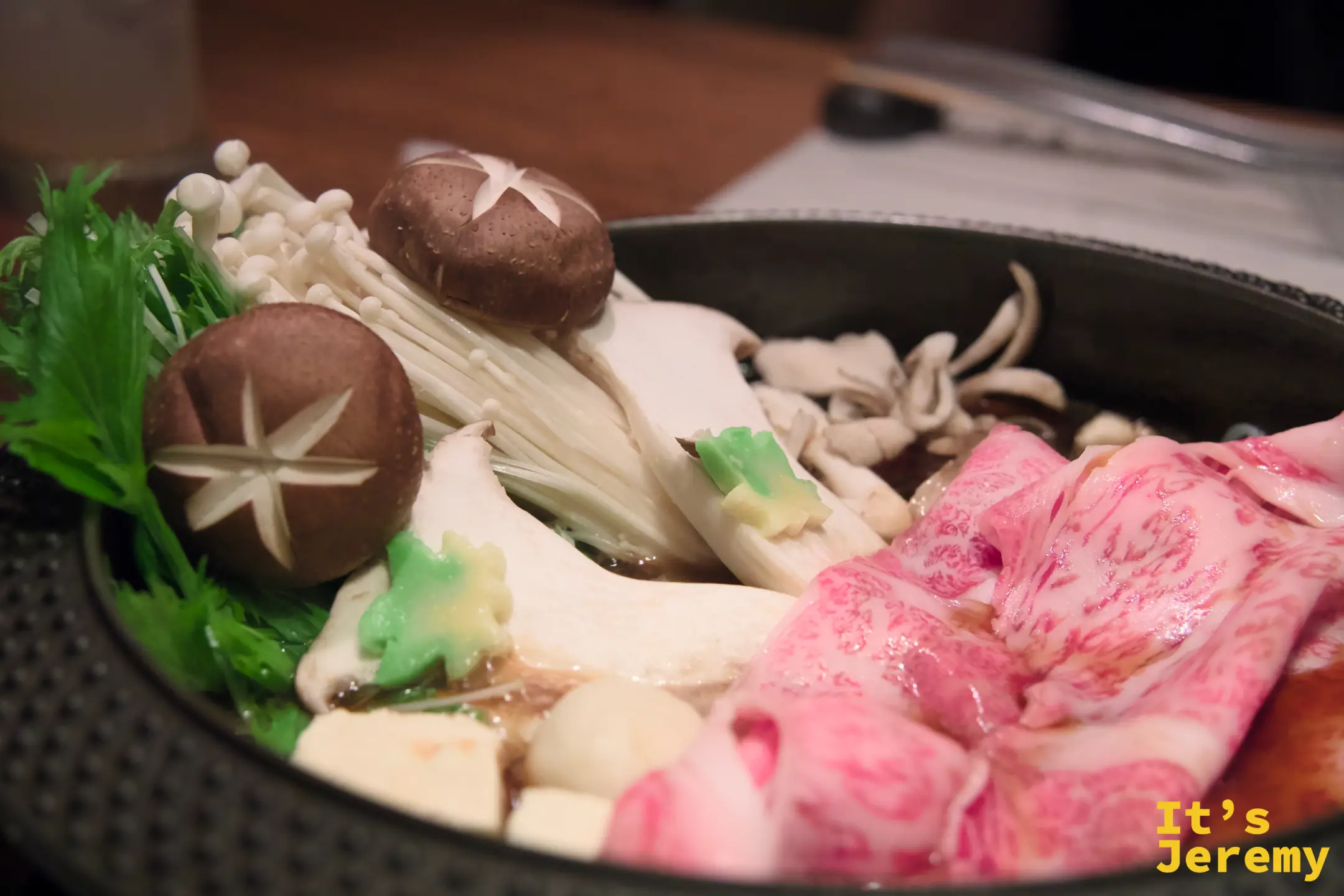 飛騨牛壽喜燒鍋端上桌,霜降肉捲與菇類蔬菜在黑色鑄鐵鍋中,食材尚未下鍋,霜降紋路在暖燈光下清晰可見