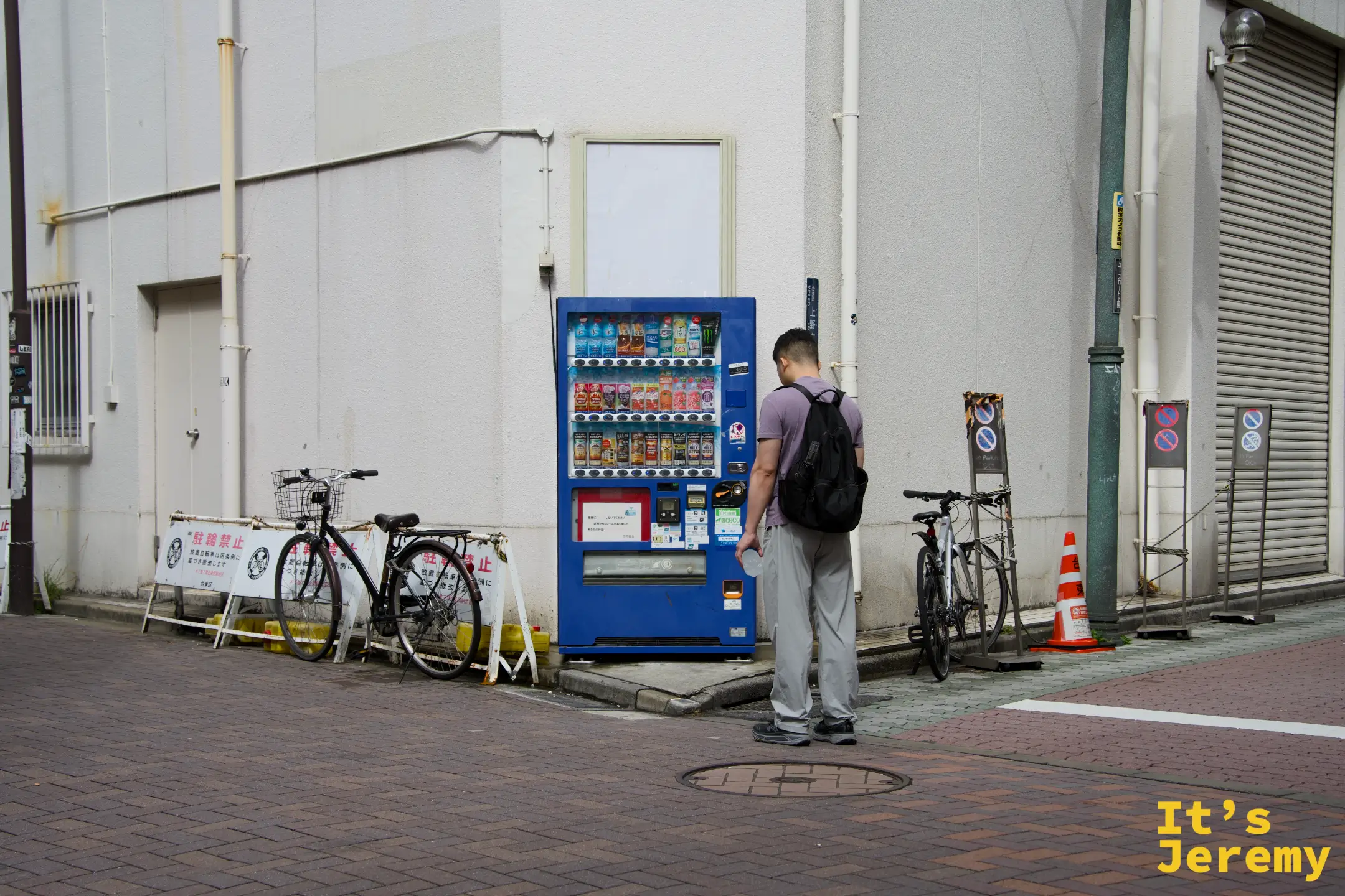 一個路人背對鏡頭站在藍色自動販賣機前選飲料,旁邊停著幾台腳踏車,是一個普通的東京街角