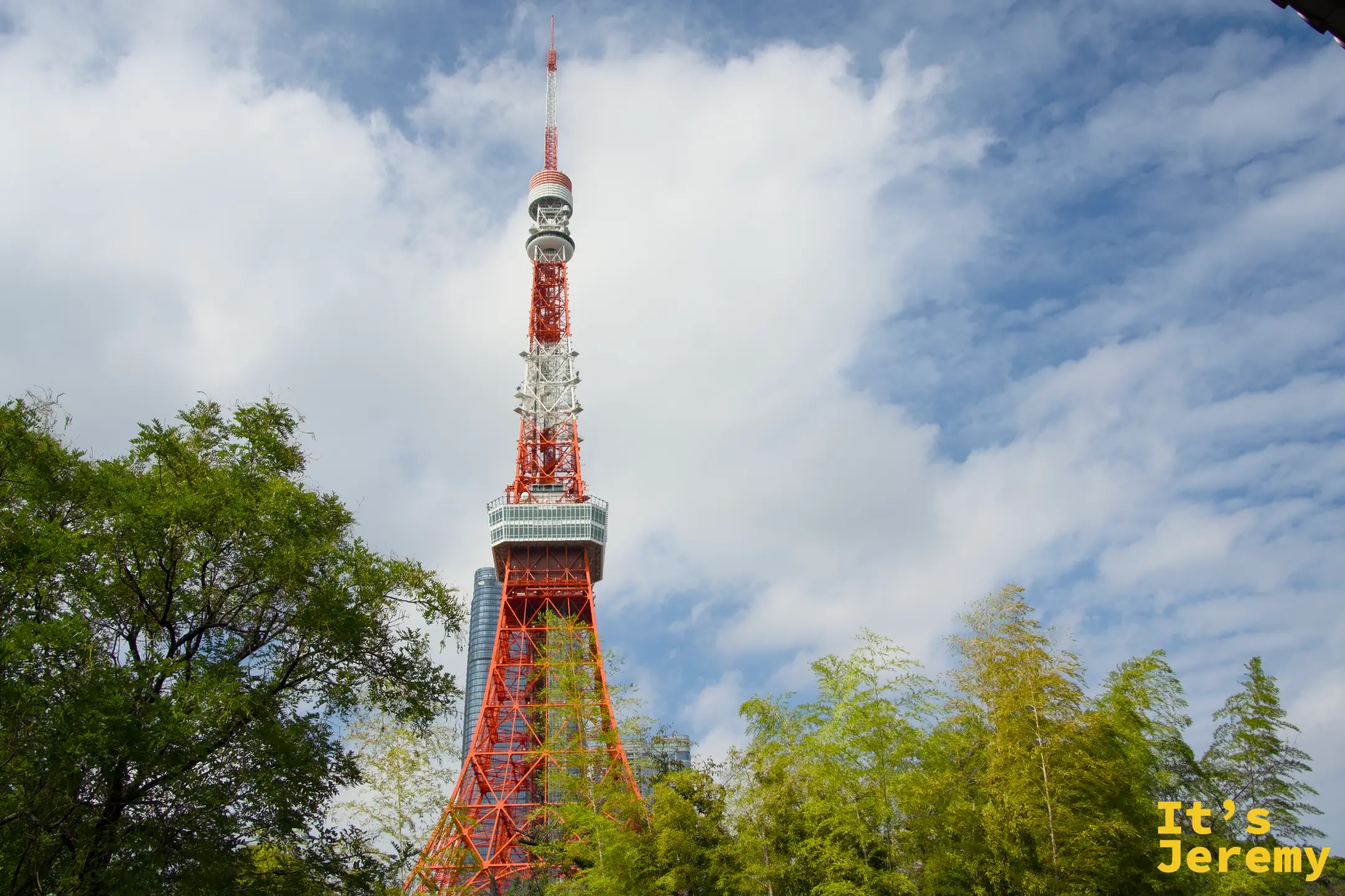 從芝公園內仰望東京鐵塔,綠樹將鐵塔局部圍起,橘紅的格架從樹梢之間拔地而起,雲朵在藍天間緩緩流動