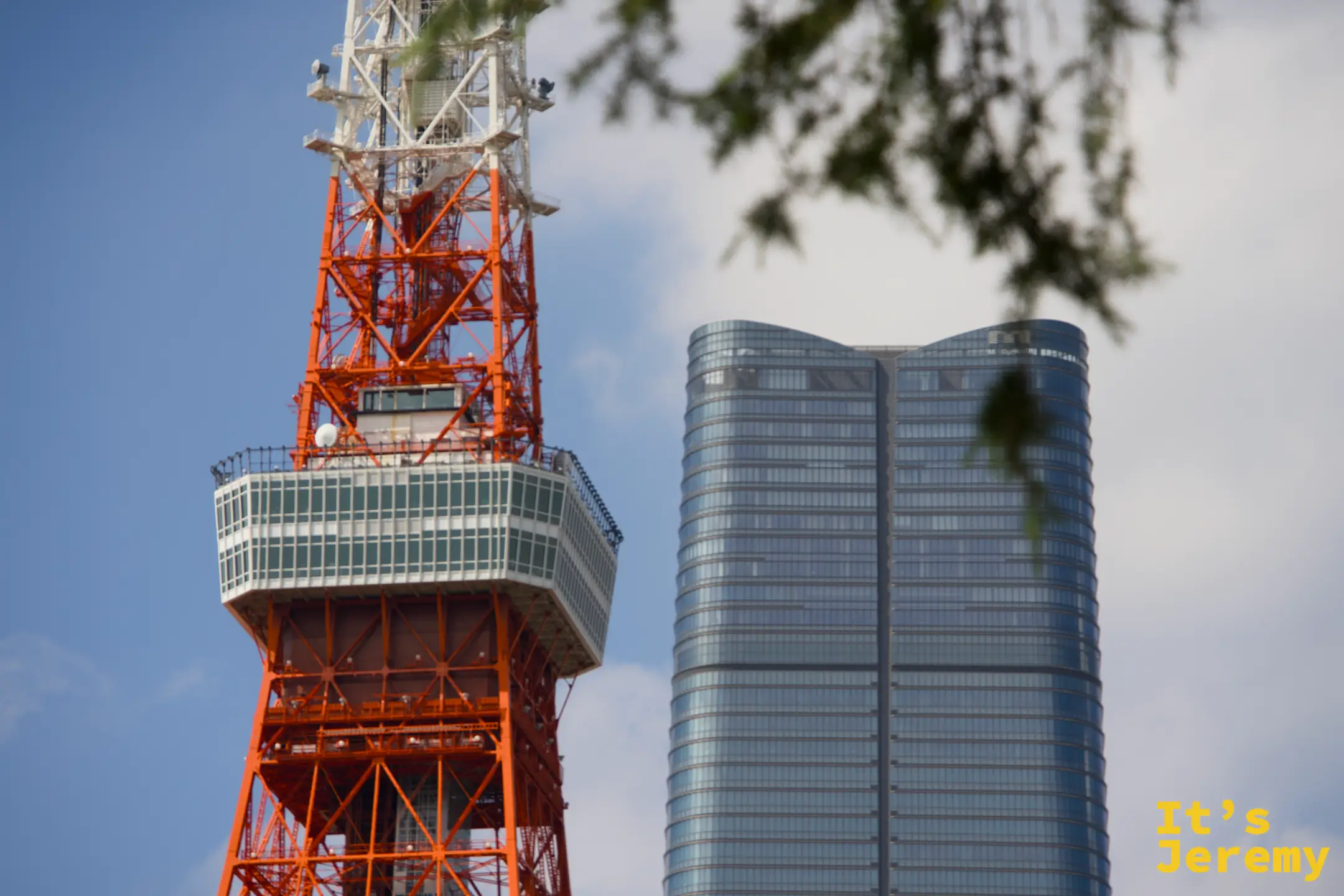 東京鐵塔的大展望台與旁邊一幢弧形玻璃帷幕高樓並排入鏡,鐵塔的橘紅格架與現代建築的冷灰玻璃形成強烈對比,前景有樹枝入鏡