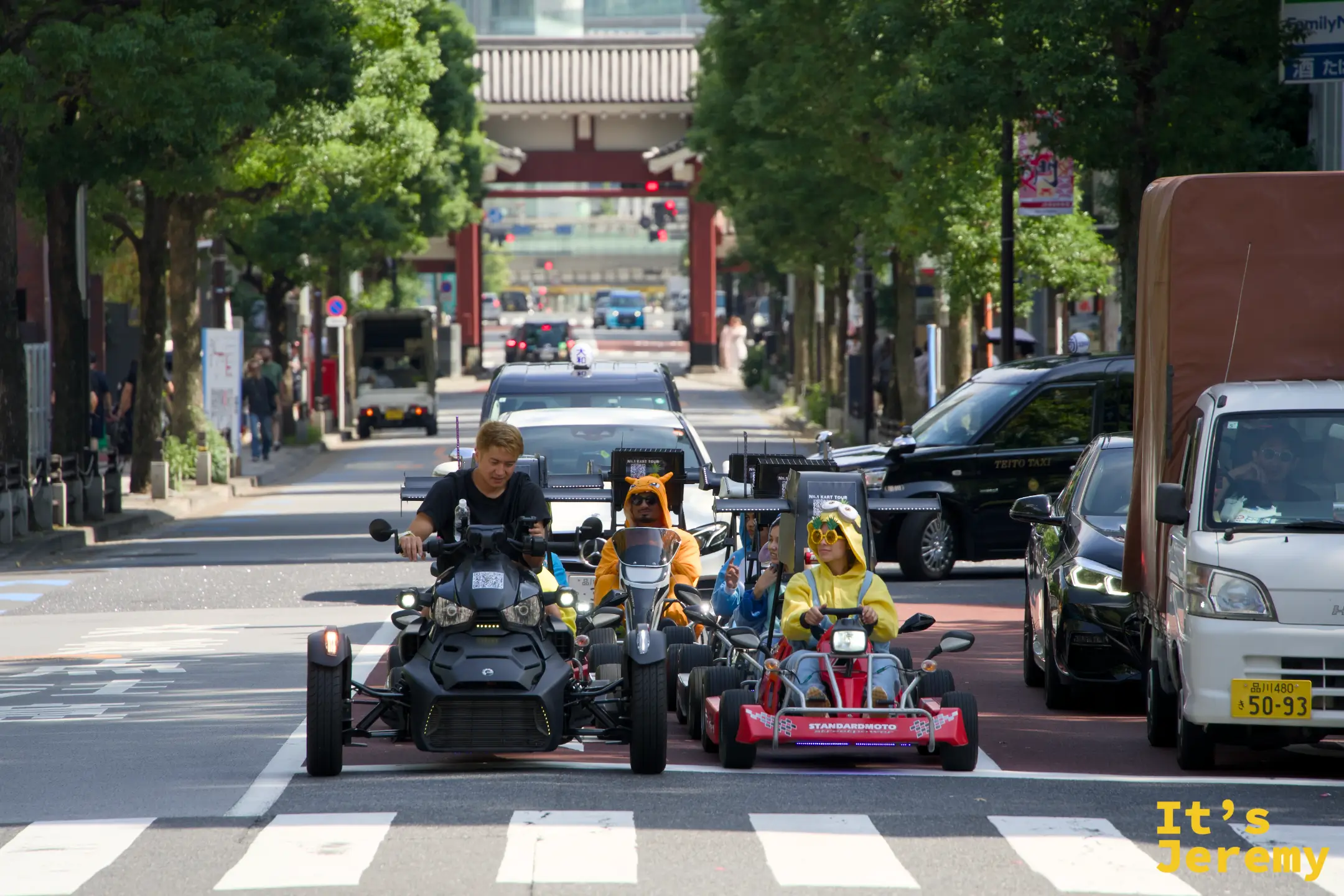 街頭車流中，幾台載著 cosplay 遊客的卡丁車混在一般車陣裡穿過路口，兩旁是行道樹與大樓