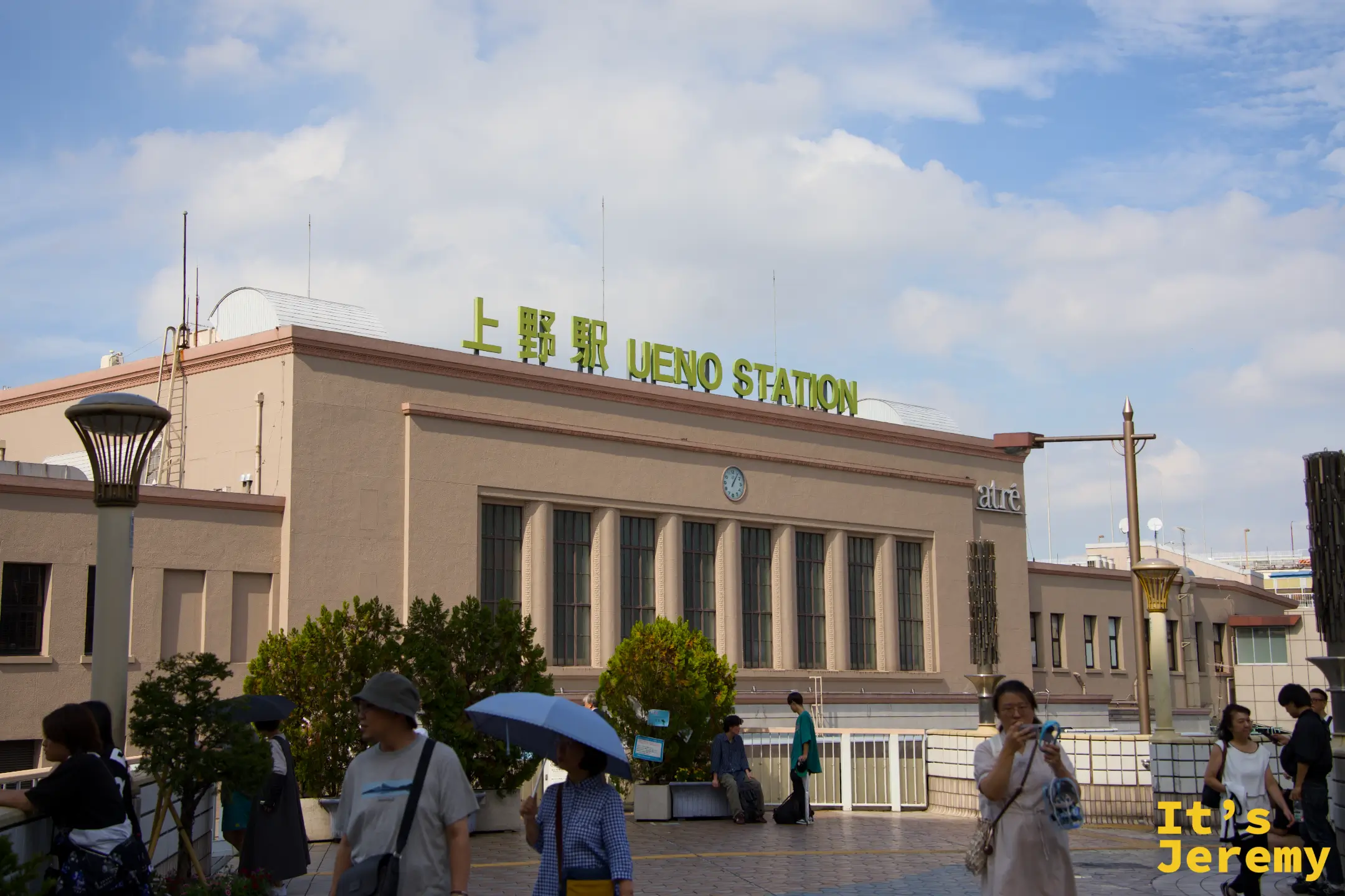 JR 上野駅外觀,「上野駅 UENO STATION」綠色大字頂在米色建築上,藍天白雲,前景是來往的旅客