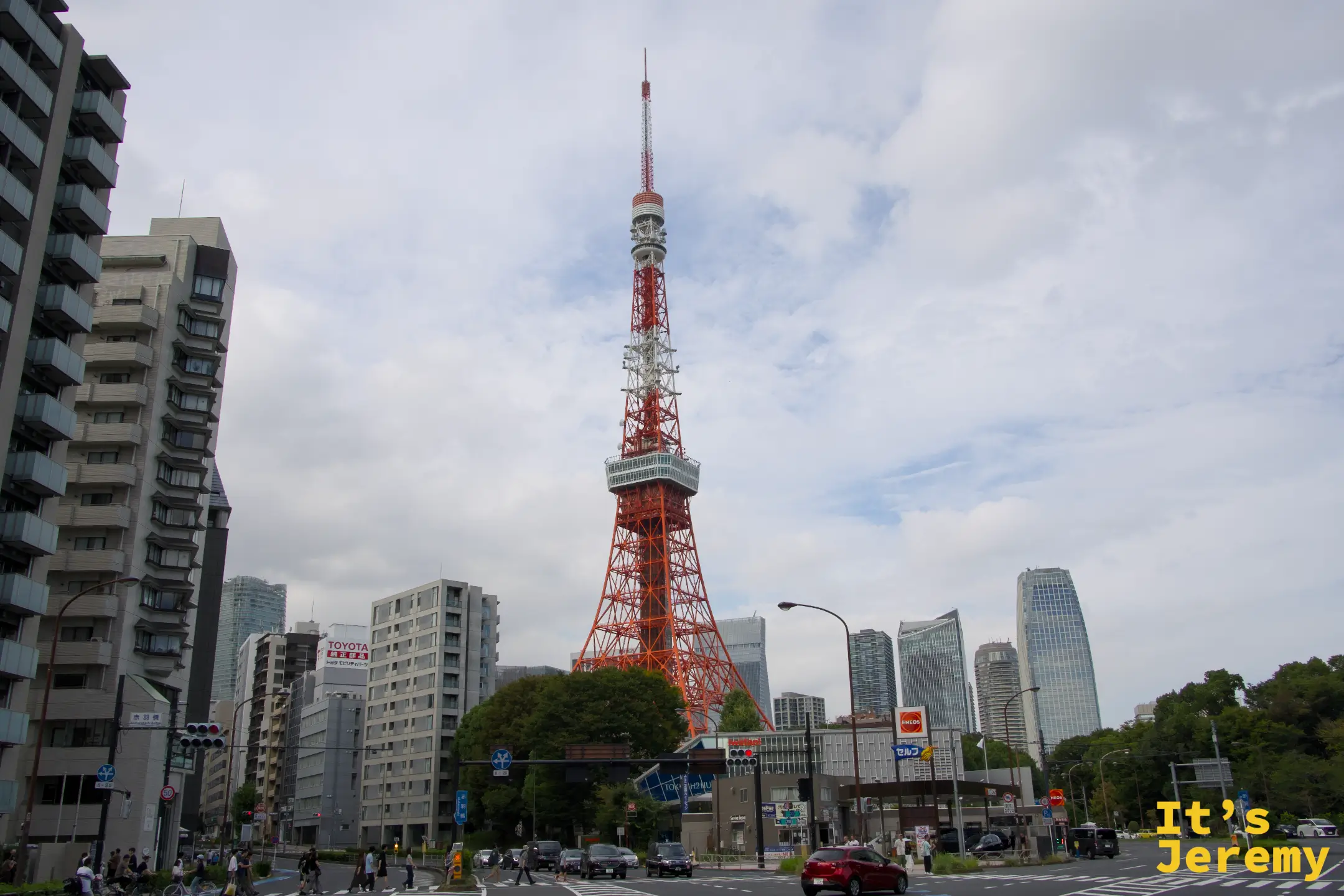 從赤羽橋交叉路口望向東京鐵塔,鐵塔矗立在城市建築群中央,橘紅色的鐵架在白色天空下格外搶眼,前景是普通的都市路口與行駛中的車輛