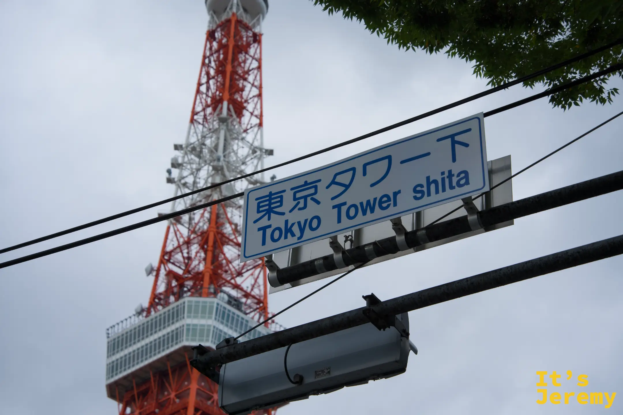 路口電線桿上的藍白路牌寫著「東京タワー下 Tokyo Tower shita」,背景是東京鐵塔的大展望台與橘紅格架,天色微陰