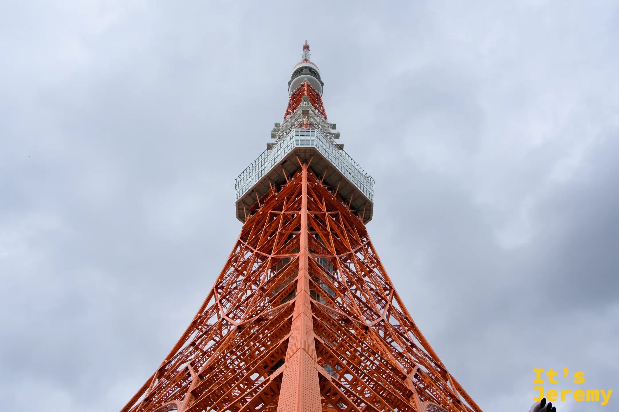 仰頭望向東京鐵塔正下方,四根主腳柱從四角往上收攏,橘紅格架在陰天的灰白天空下顯得格外有份量,大展望台在畫面中段懸空而立