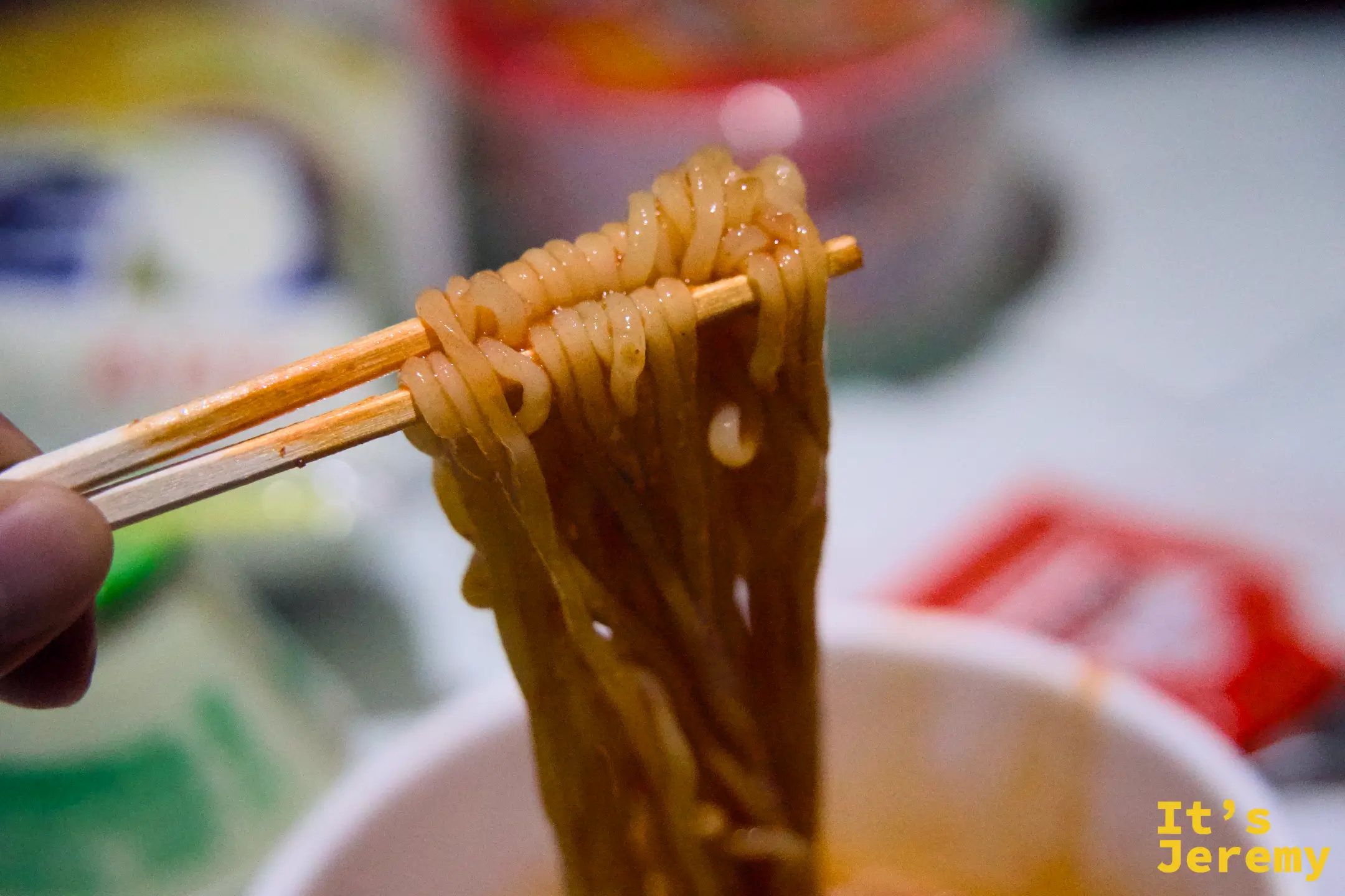 用筷子夾起一束麵條，背景是飯店房間桌面，泡麵包裝散落其間