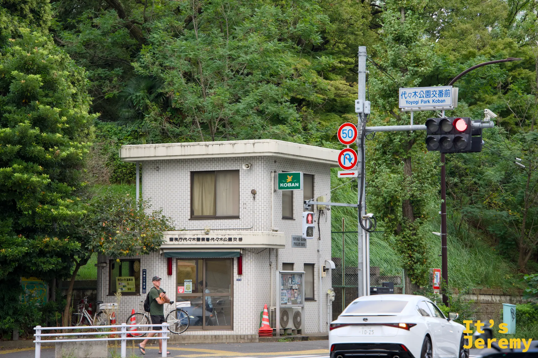 代々木公園交差點,角落是白色交番建築,旁邊路牌寫著「代々木公園交差点」,周圍樹木茂密,白色轎車靜靜停在路邊