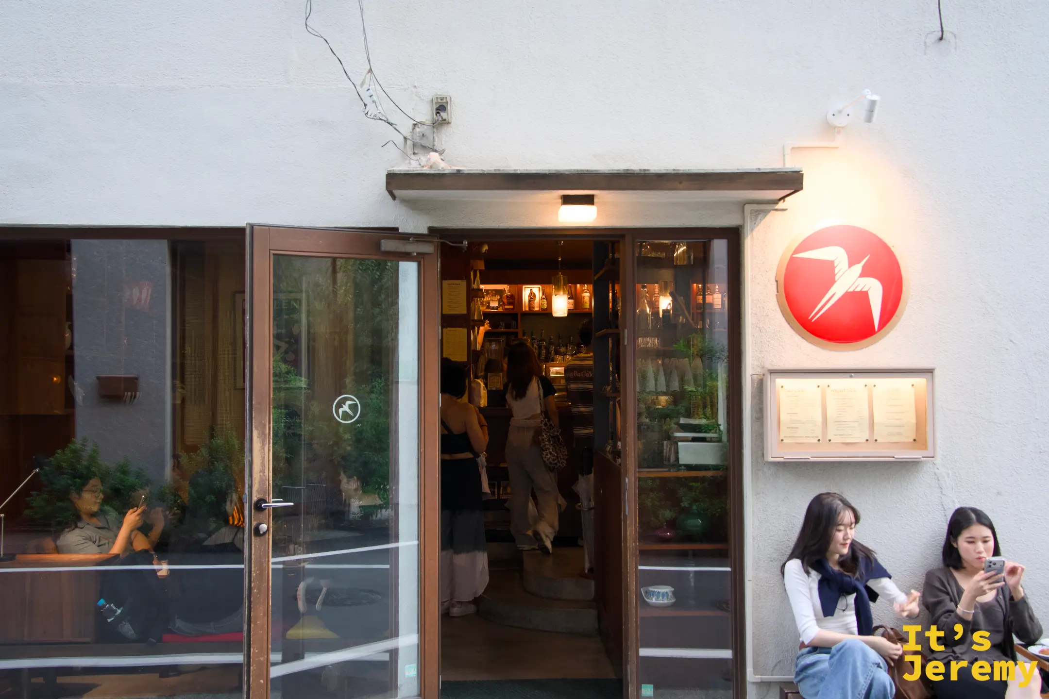 Fuglen Tokyo 入口近景,橙色燕子 logo 圓牌,玻璃門半開,裡頭一片暖黃燈光