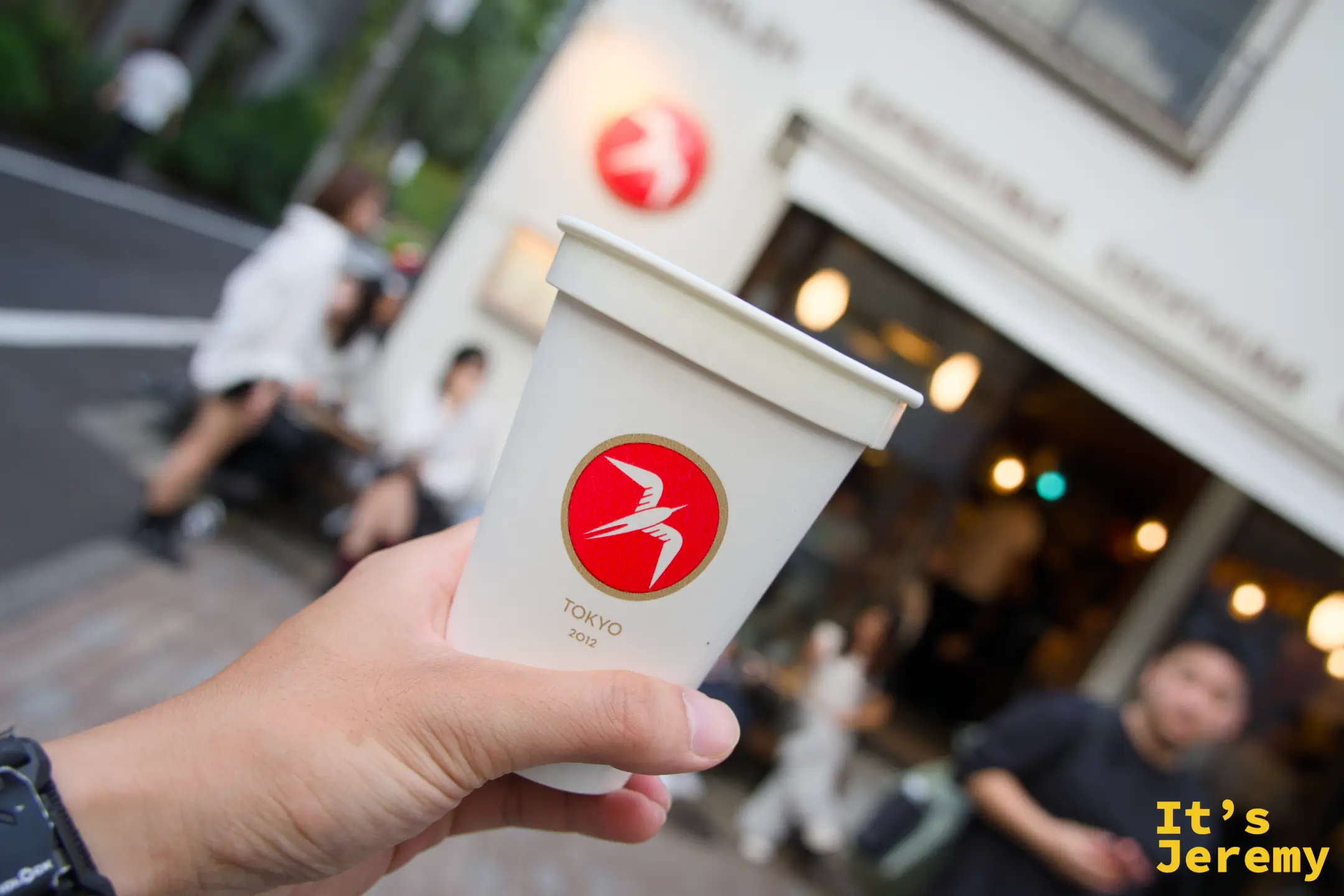 手持 Fuglen TOKYO 2012 紙杯，背景是店門外的長椅與暖黃燈光