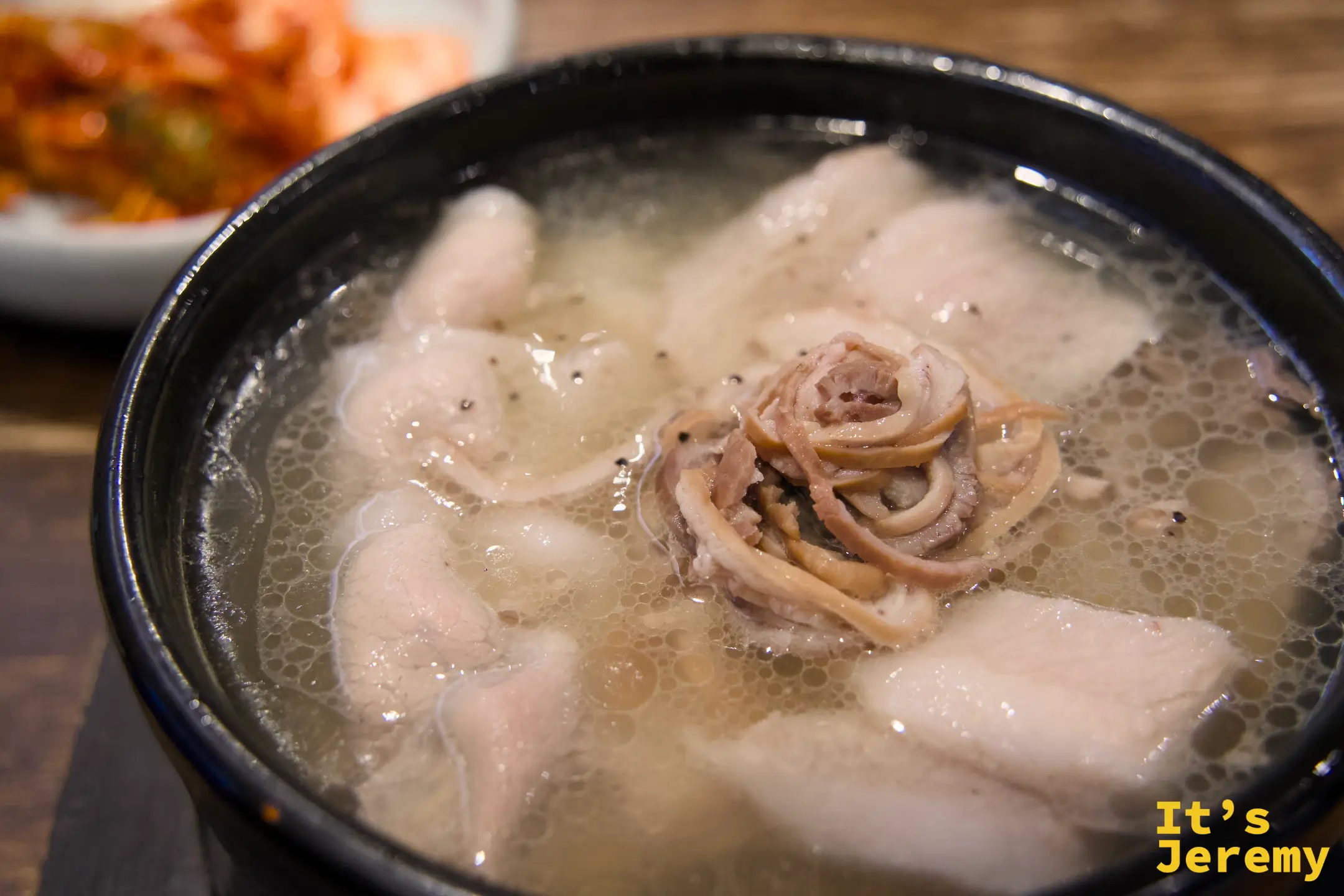 釜山式豬肉湯飯近景，清亮湯底中漂浮著厚切豬肉片與捲曲的豬橫膈膜，後方是泡菜小碟