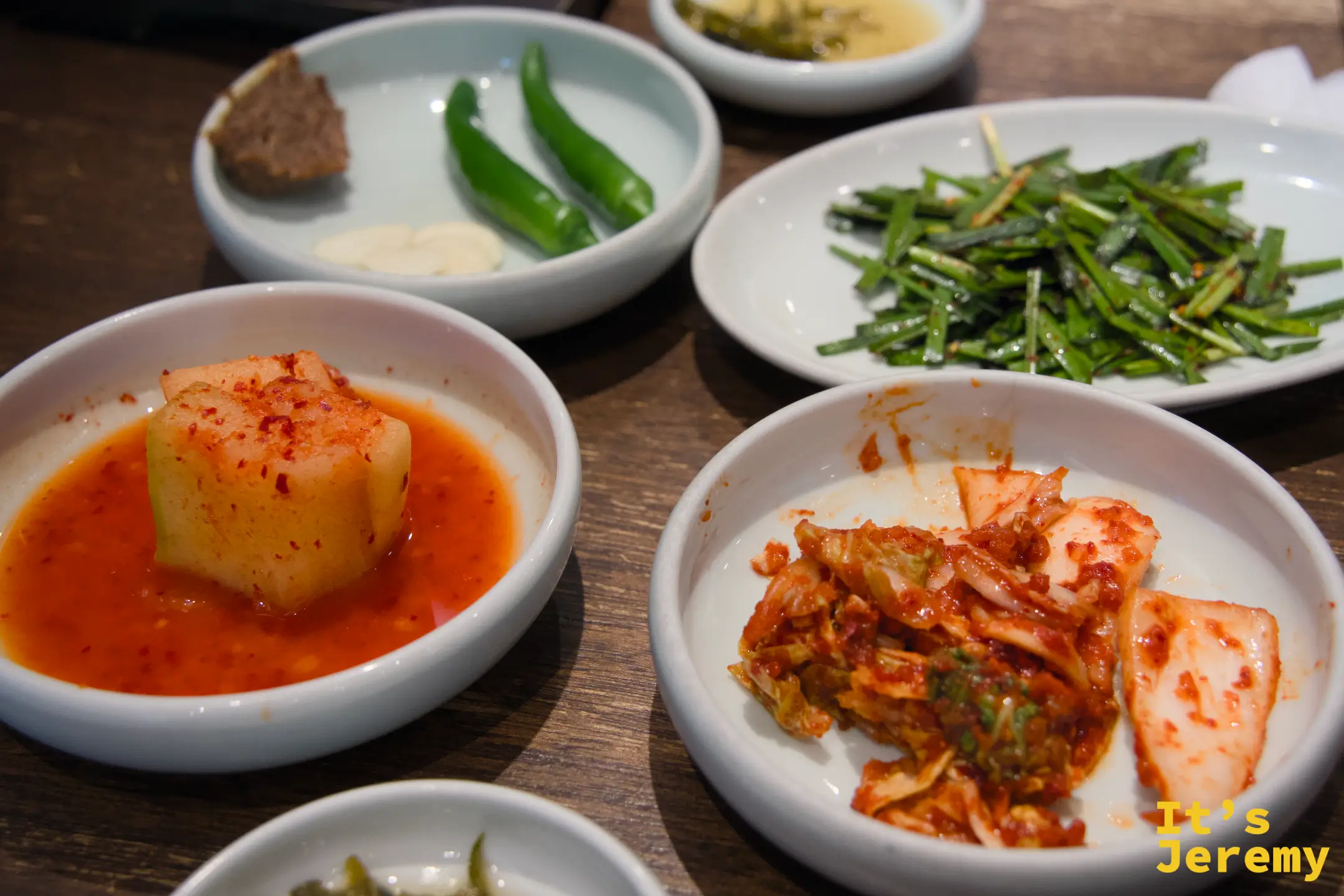 餐桌上豐盛的小菜配菜：方塊蘿蔔泡菜、白菜泡菜、青辣椒配醬與青蔥沙拉，色彩鮮豔對比