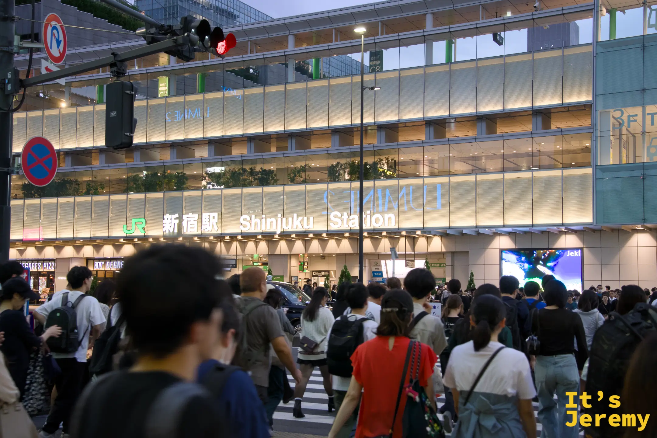 傍晚的 JR 新宿駅南口,LUMINE 玻璃帷幕大樓燈光全亮,「新宿駅 Shinjuku Station」招牌打燈,路口人群正在穿越斑馬線