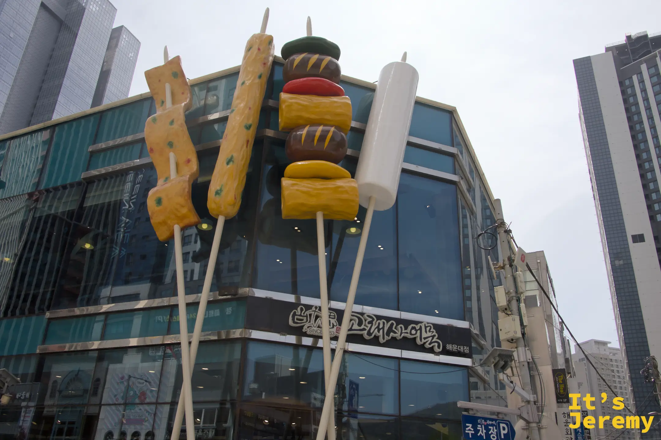 從路口望向古來思魚板海雲台店，三根巨型魚板串矗立街角，海雲台鬧區高樓為背景