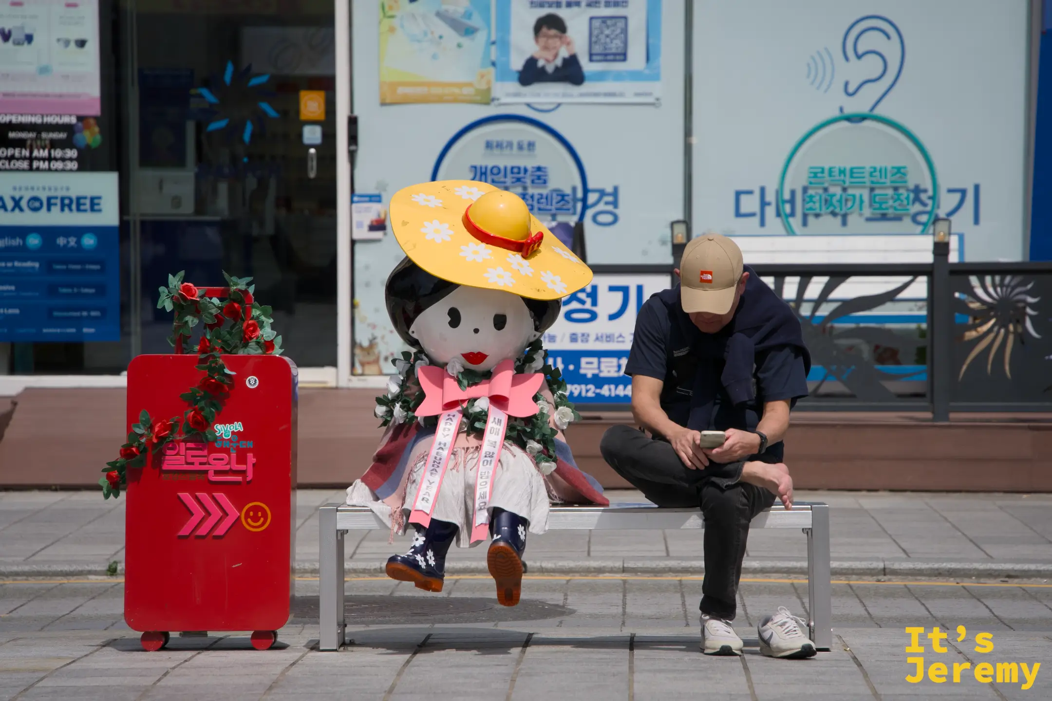 海雲台街頭,穿著傳統韓服、戴著黃色大草帽的可愛偶人裝置坐在長椅上,旁邊的男子低頭滑手機;這種人與裝置共存的街頭畫面,是釜山日常裡意外好拍的素材