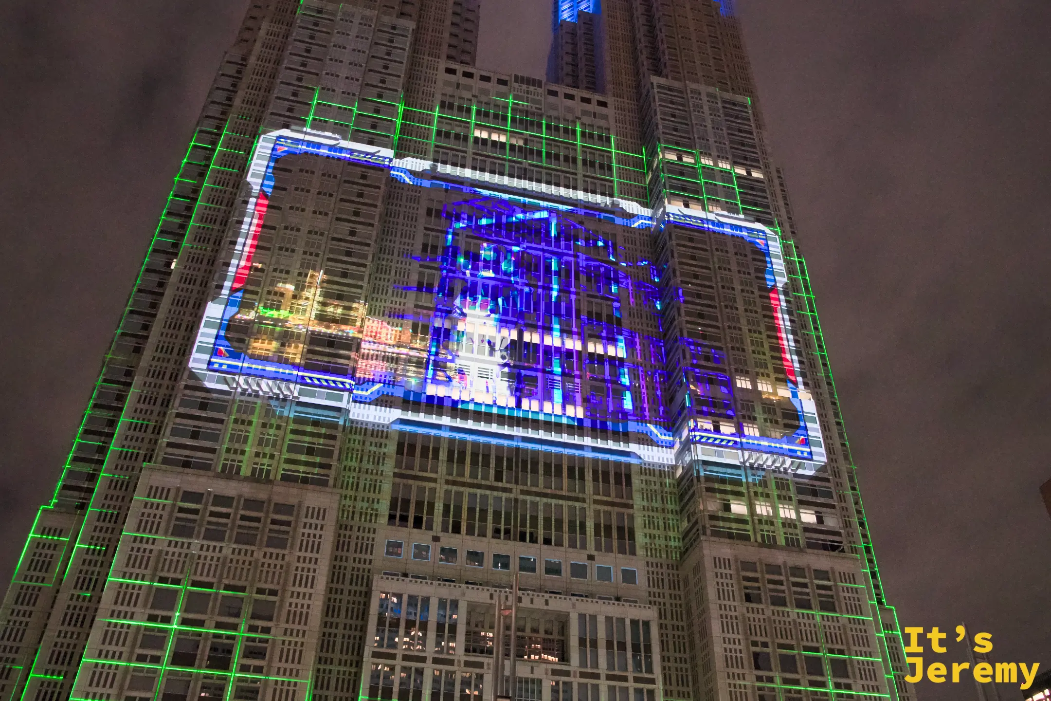東京都廳大樓全景，整棟外牆覆蓋藍色科技感框線，配以綠色格狀光網，入夜後如同被電路板包住