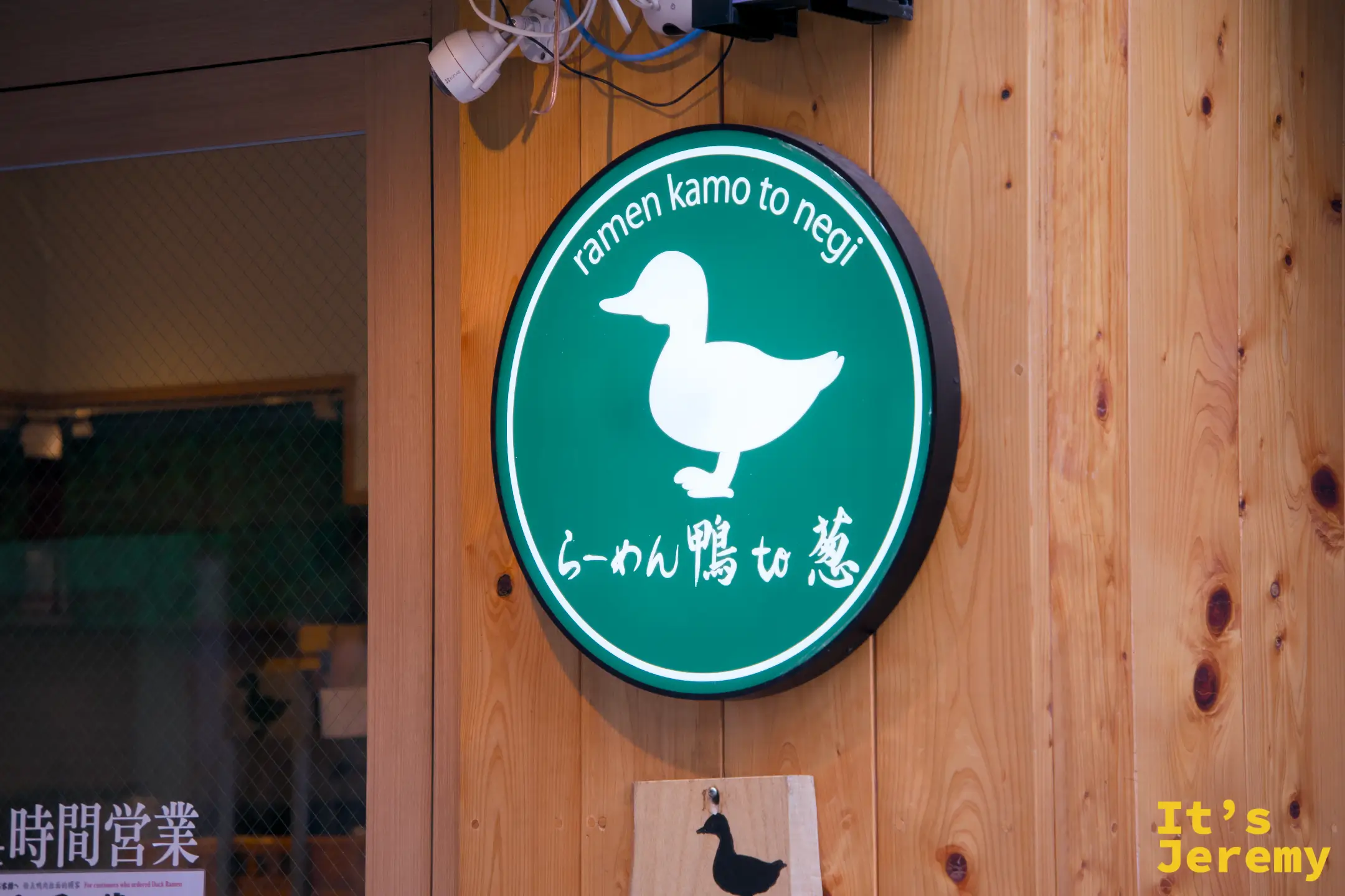 店門圓形logo燈箱，白色鴨子剪影在綠色底上，下方英文「ramen kamo to negi」
