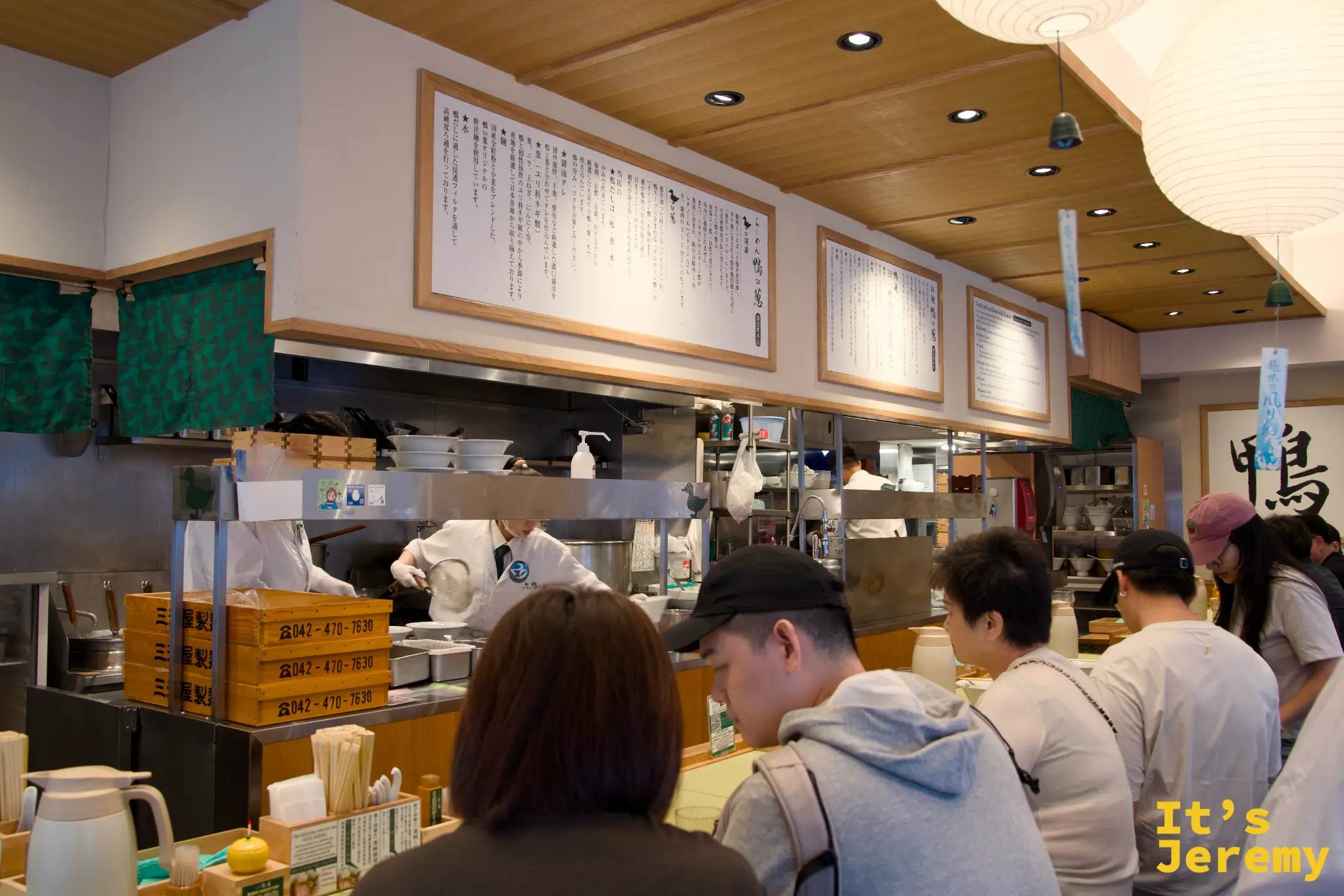 店內吧台座位，多位客人面對廚房坐下，廚師忙碌，牆上說明文字框，右側掛著「鴨」字書法