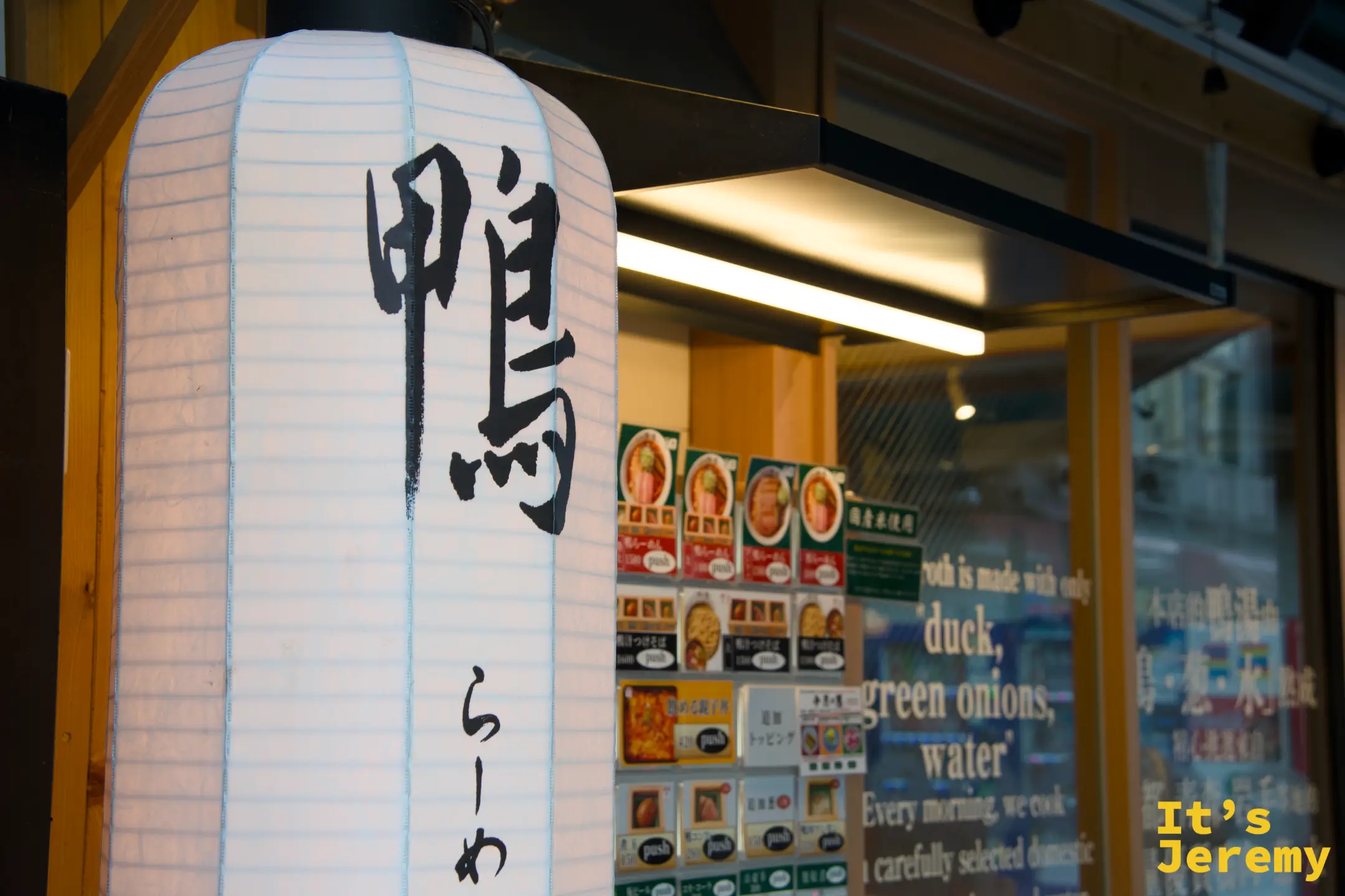 店門口懸掛的白色燈籠，寫著「鴨らーめん」，後方是自動售票機菜單