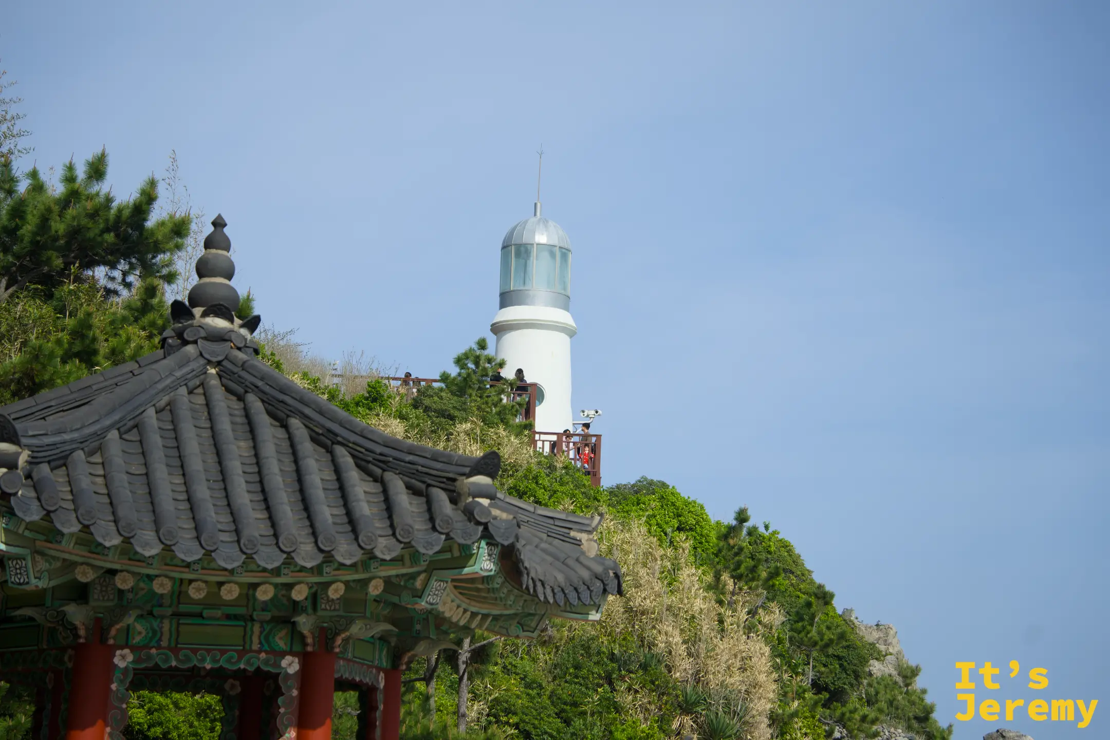 前景是傳統韓式亭子的飛簷，背景山丘上是白色燈塔，兩者的年代對比鮮明