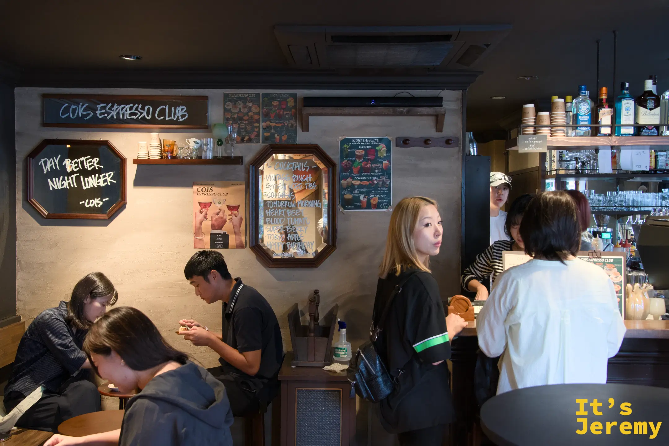 店內氛圍:客人在暖光下落座,「DAY BETTER NIGHT LONGER」手寫黑板與調酒菜單掛在同一面牆上