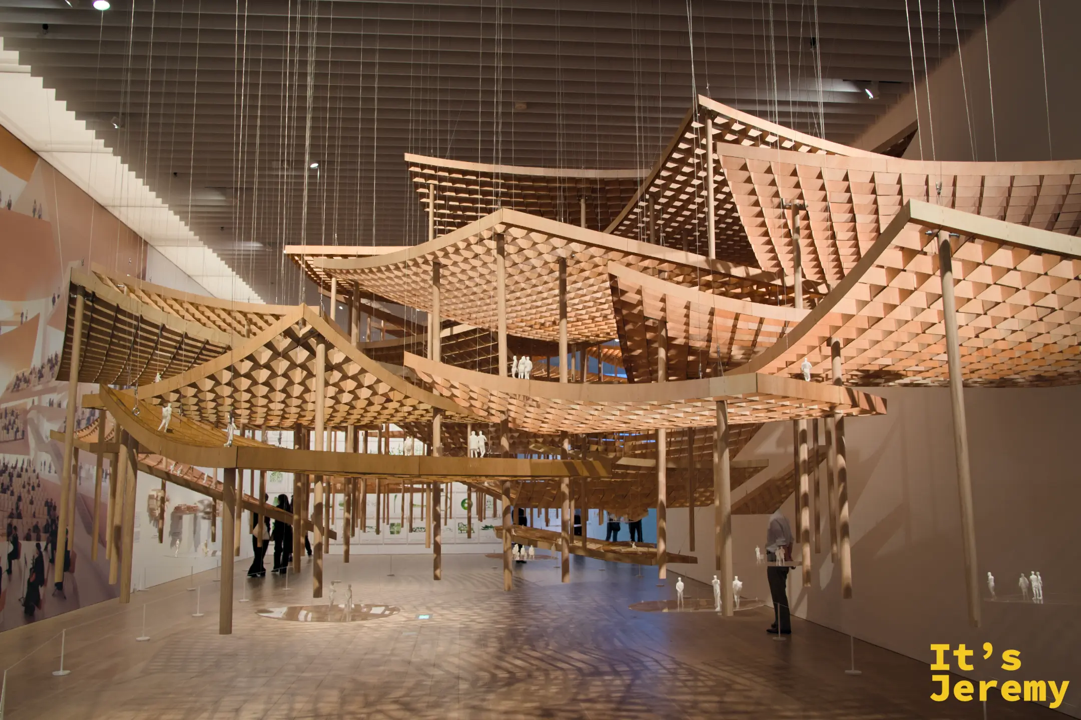 懸吊於展廳中央的波浪形大型木造建築模型，靠細鋼索從天花板垂落，白色人物模型點綴其間