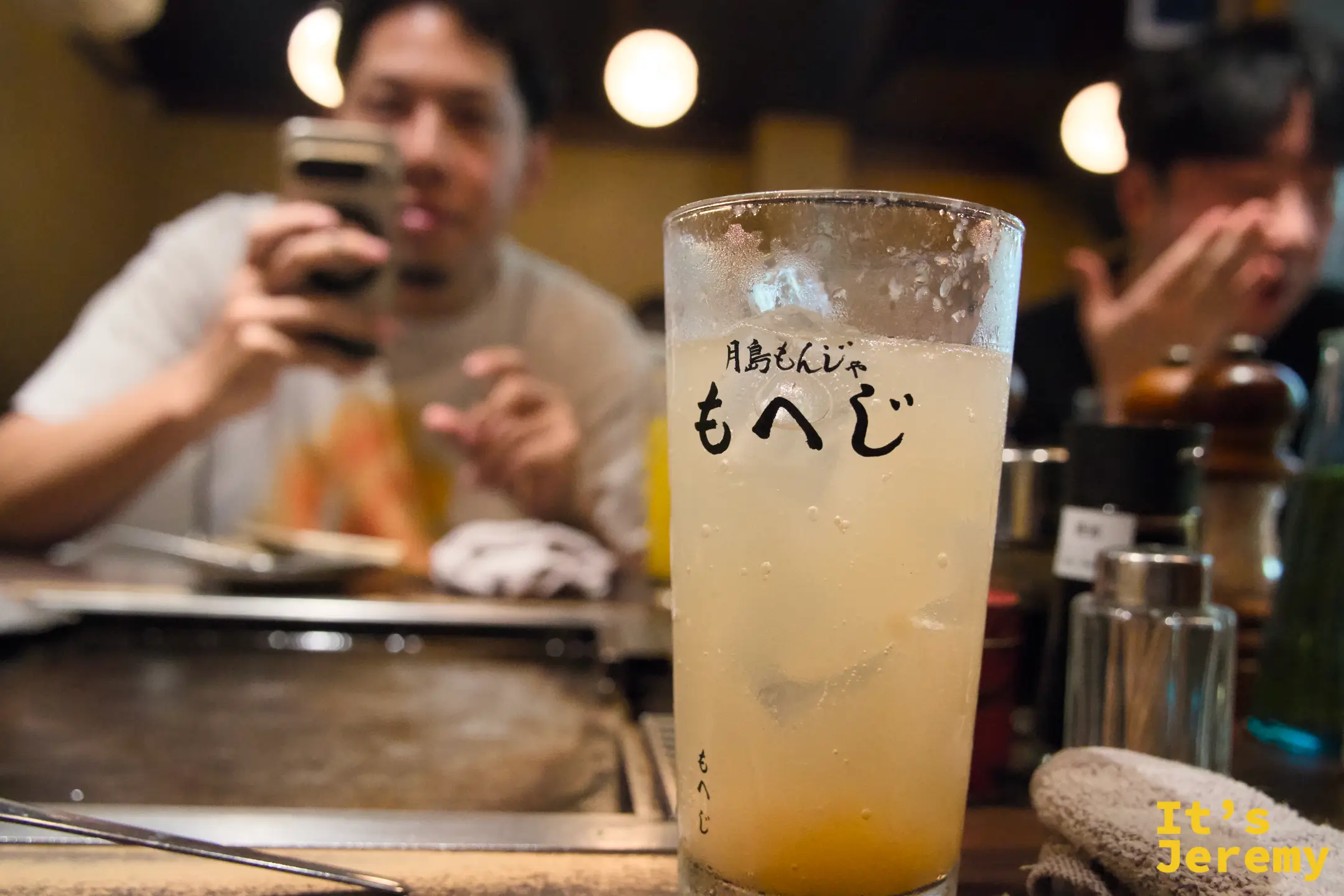 もへじ 店名玻璃杯裝的黃橙色飲品，背景是冒煙的鐵板與食材