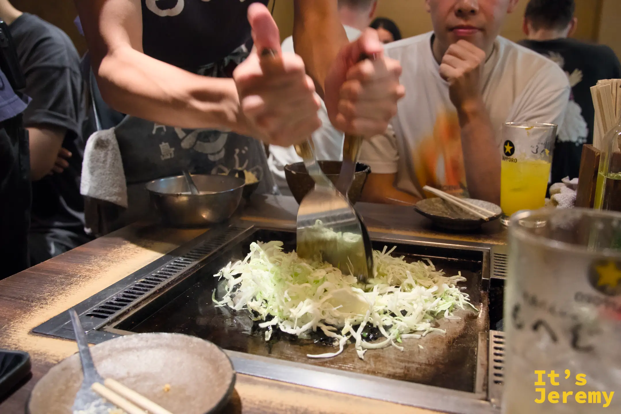 店員用兩把鐵鏟在鐵板上翻炒高麗菜絲，鋪平準備迎接文字燒麵糊