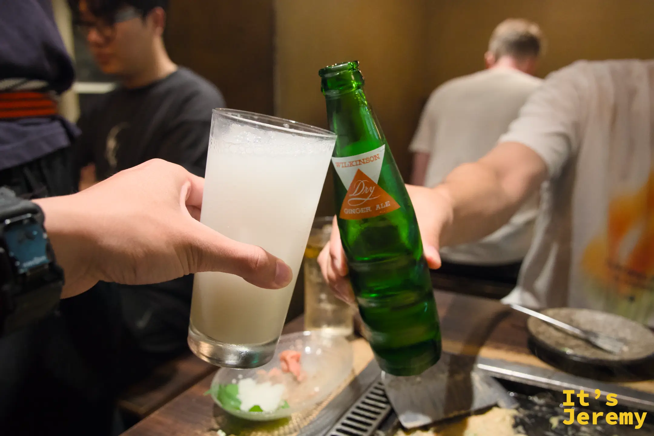舉起一杯白色混濁飲品，對面是 Wilkinson Dry Ginger Ale 綠色玻璃瓶，周圍是冒著熱氣的鐵板