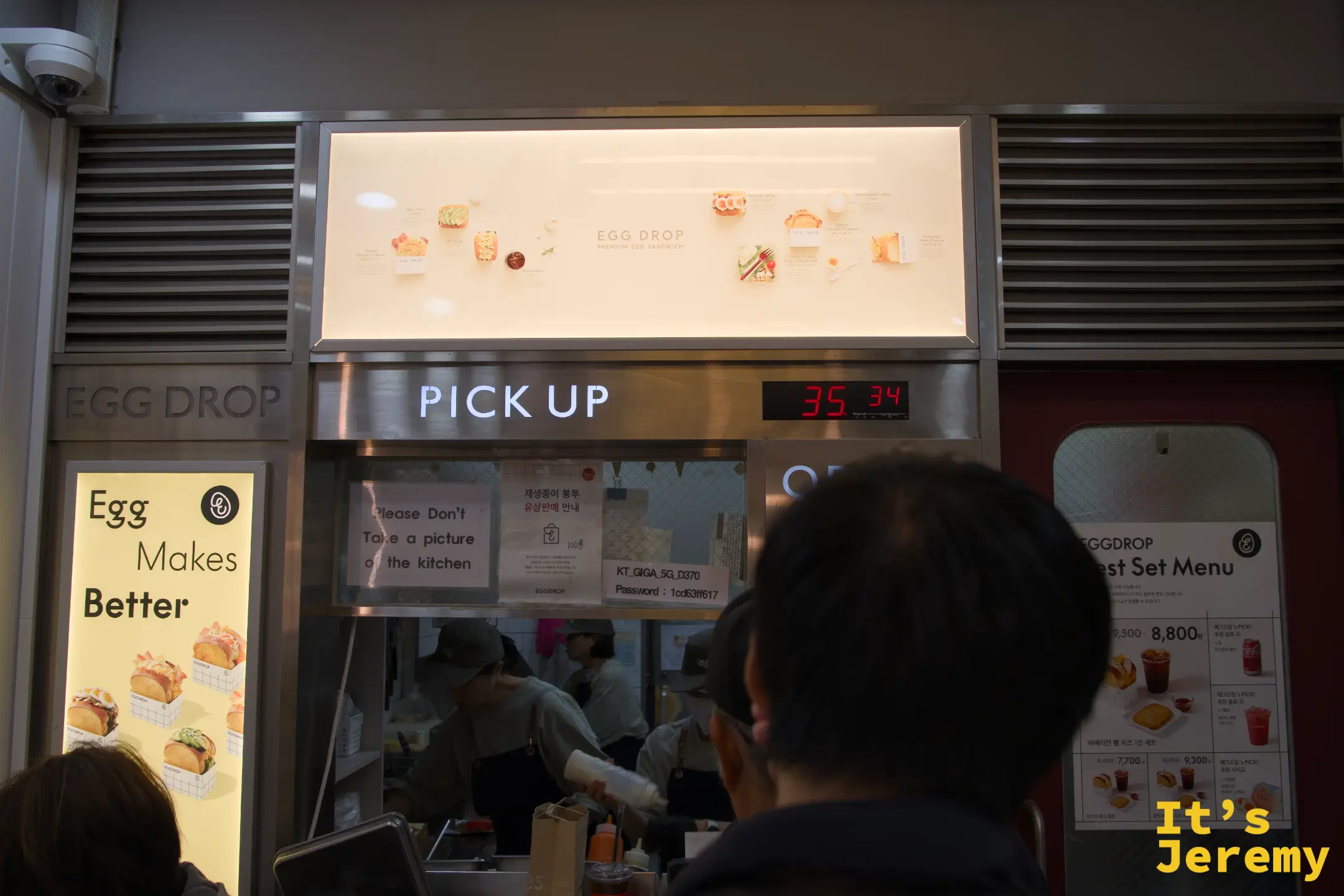 店內 PICK UP 區，電子看板顯示目前叫號，背後廚房一直在備餐