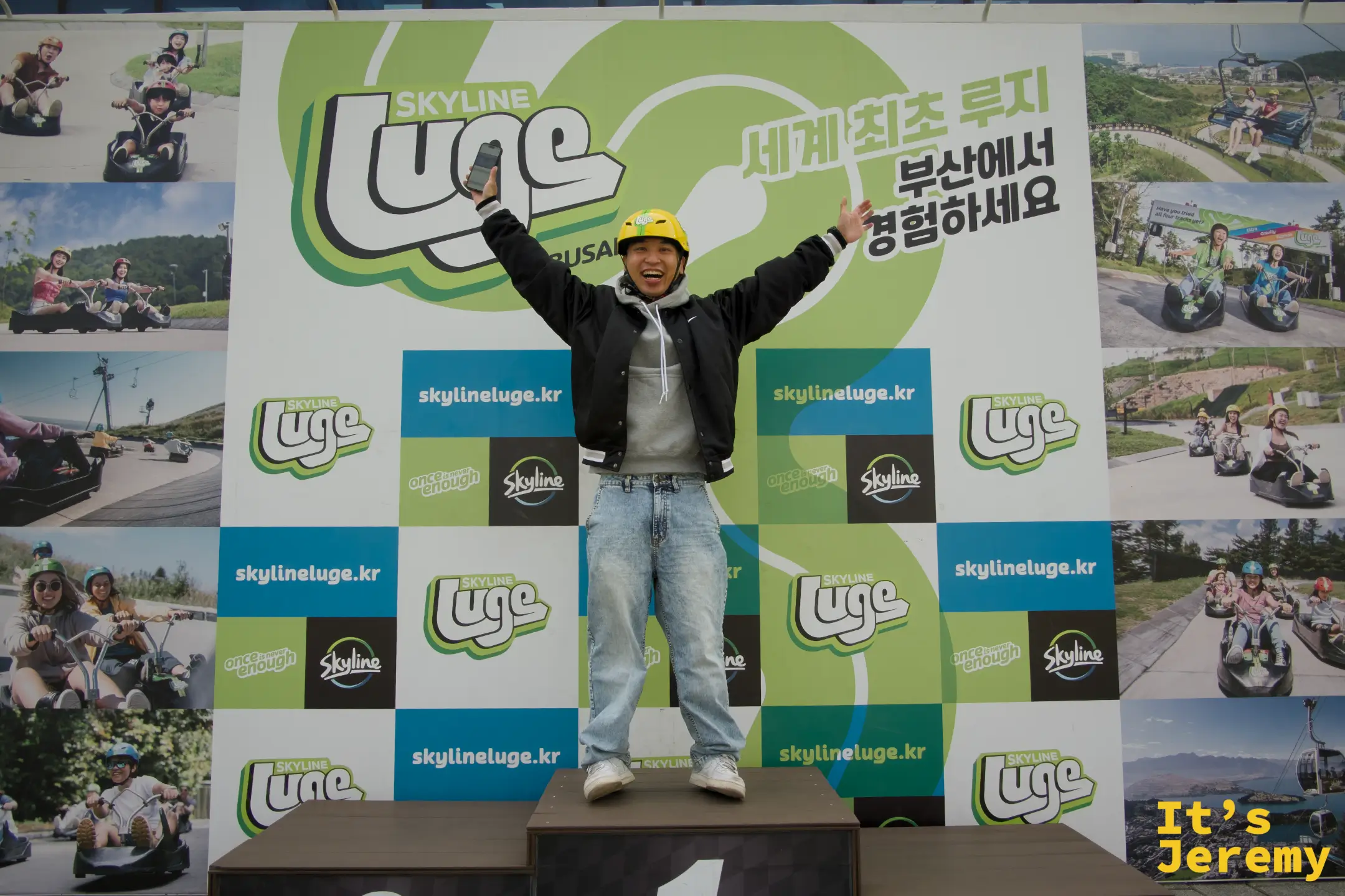 Jeremy 戴著黃色安全帽站在頒獎台第一名位置，雙手舉高慶祝，背後是 Skyline Luge 品牌大牆與「세계 최초 루지 부산에서 경험하세요」字樣