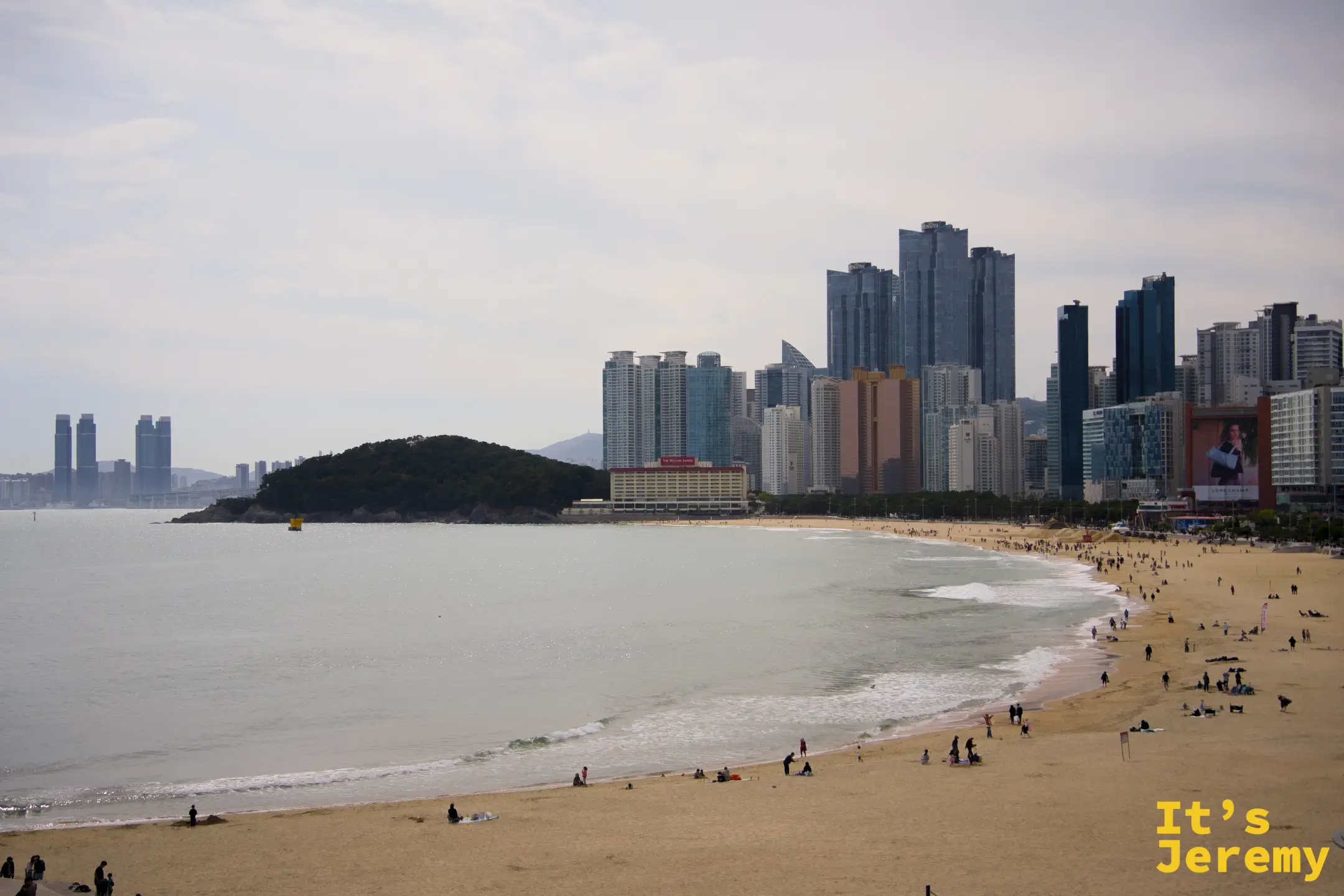 海雲台全沙灘弧線，從防波堤望向冬柏島方向，高樓與海灘交疊