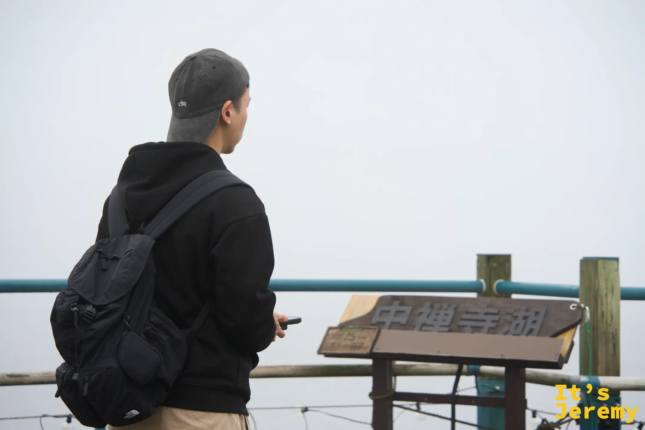 背包旅人站在「中禪寺湖」木製標示牌前，望向白茫茫的湖面