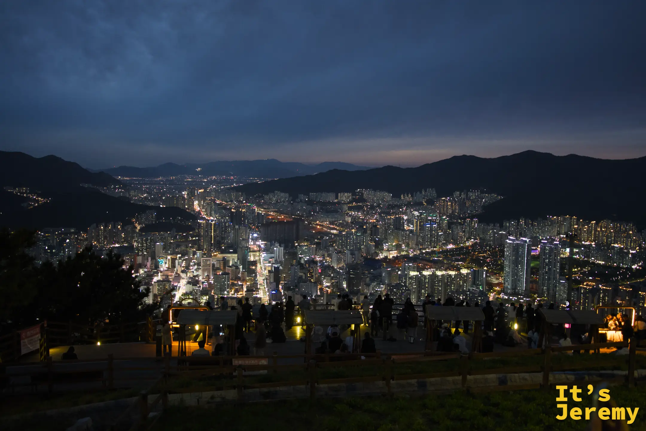 日落剛過，遊客在木製觀景台上坐下，山谷間的釜山市區燈火逐漸亮起