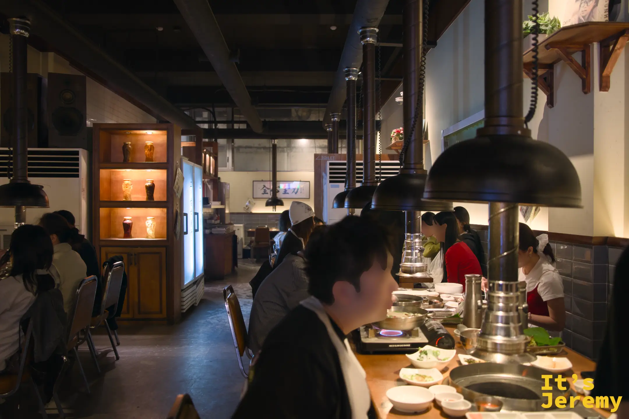 餐廳內部氛圍：黑鐵吊燈、木桌、排氣管道，工業復古調性在暖燈下顯得格外溫潤