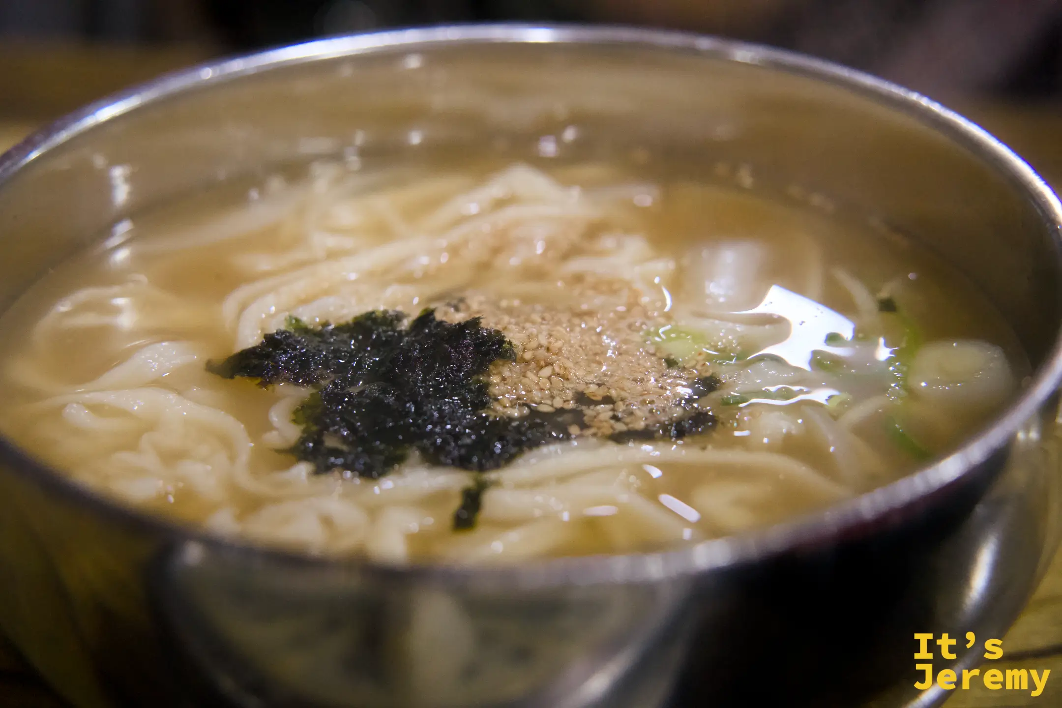 칼국수的近景：清澈骨湯、寬厚手切麵條，頂上撒著海苔碎與白芝麻