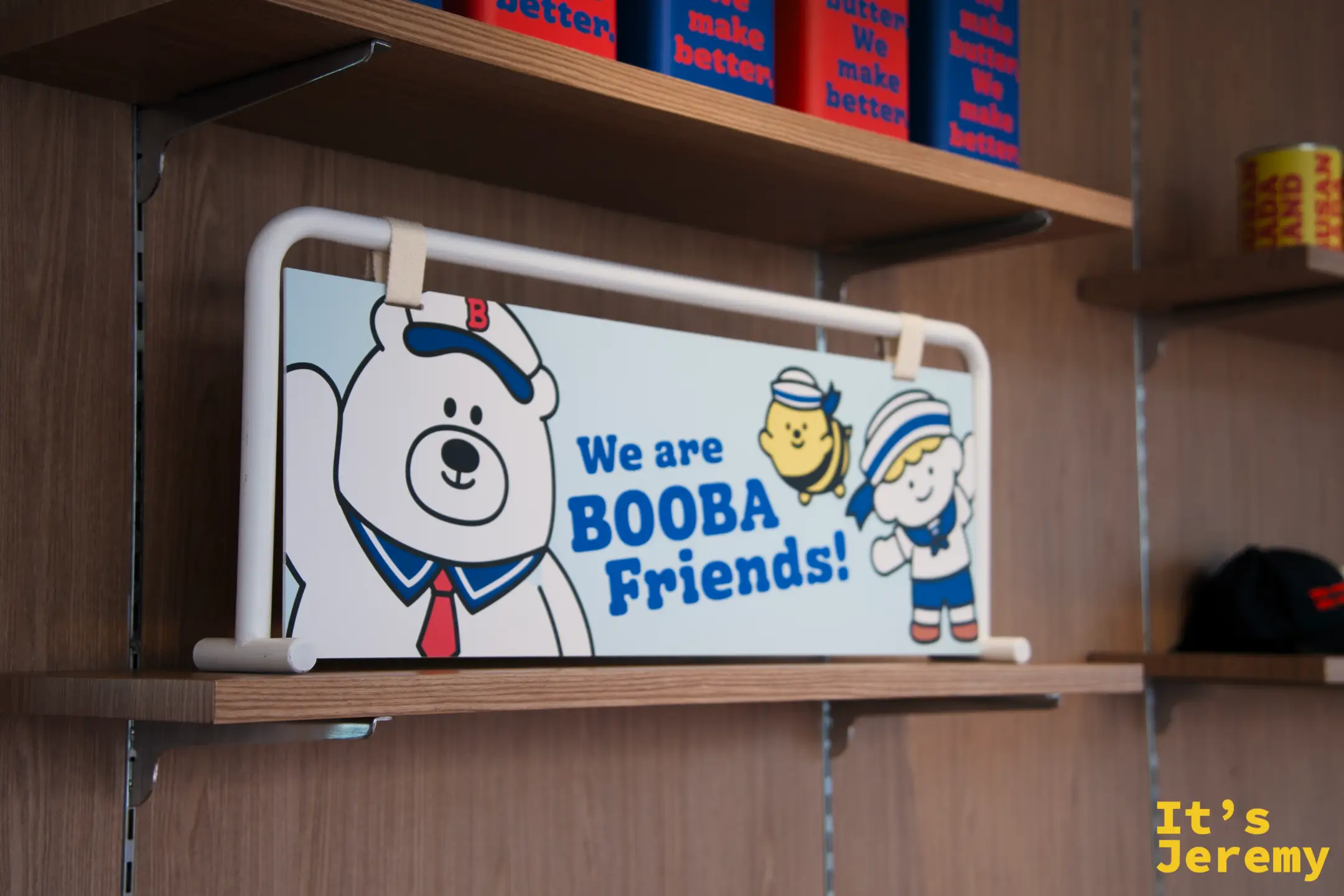 店內木架上的「We are BOOBA Friends!」角色插畫板，BOOBA 熊、蜜蜂和水手女孩三人齊聚，架上排著藍橘配色商品罐