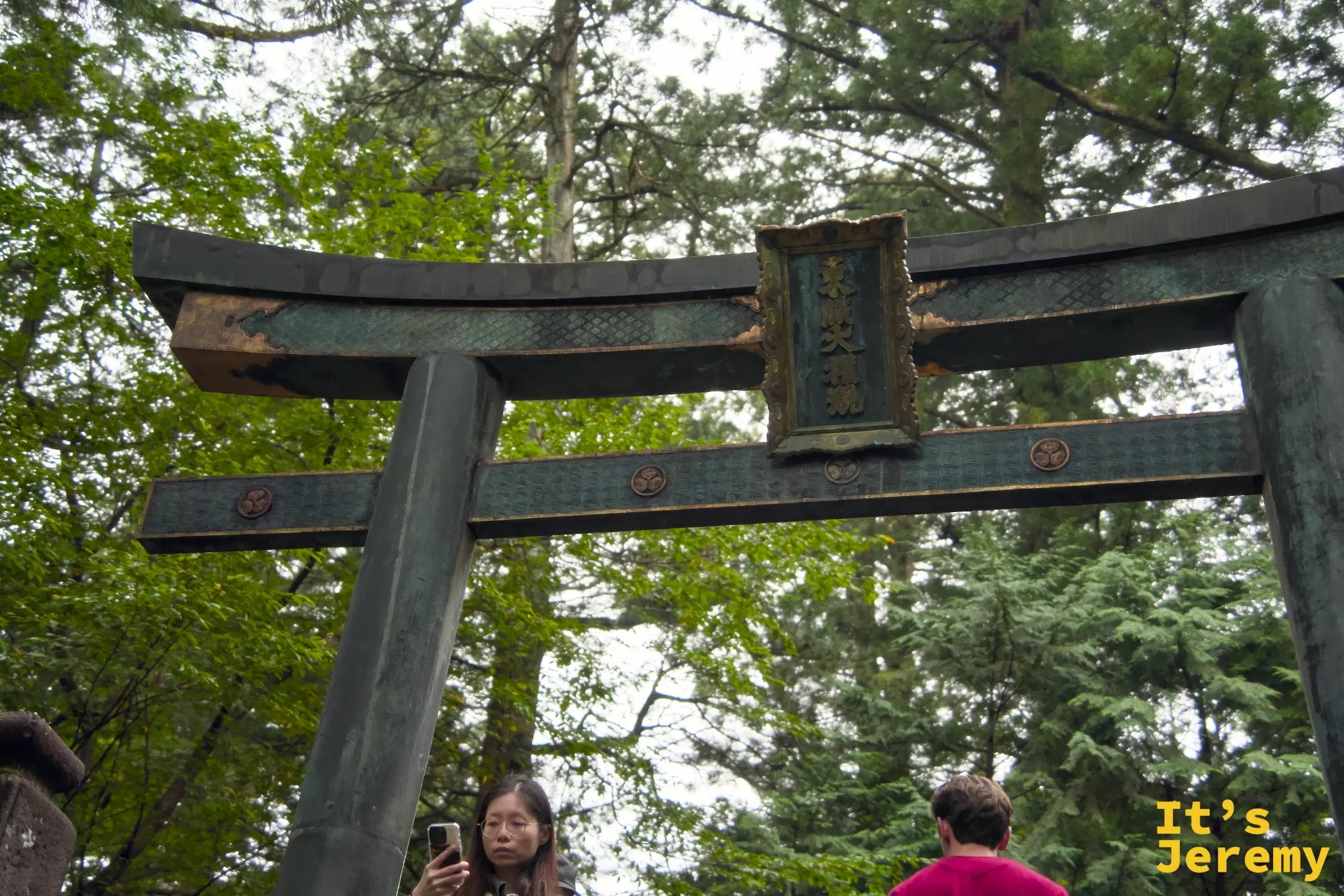 遊客在青銅鳥居下方仰拍留念，鳥居巨大的曲線橫跨頭頂