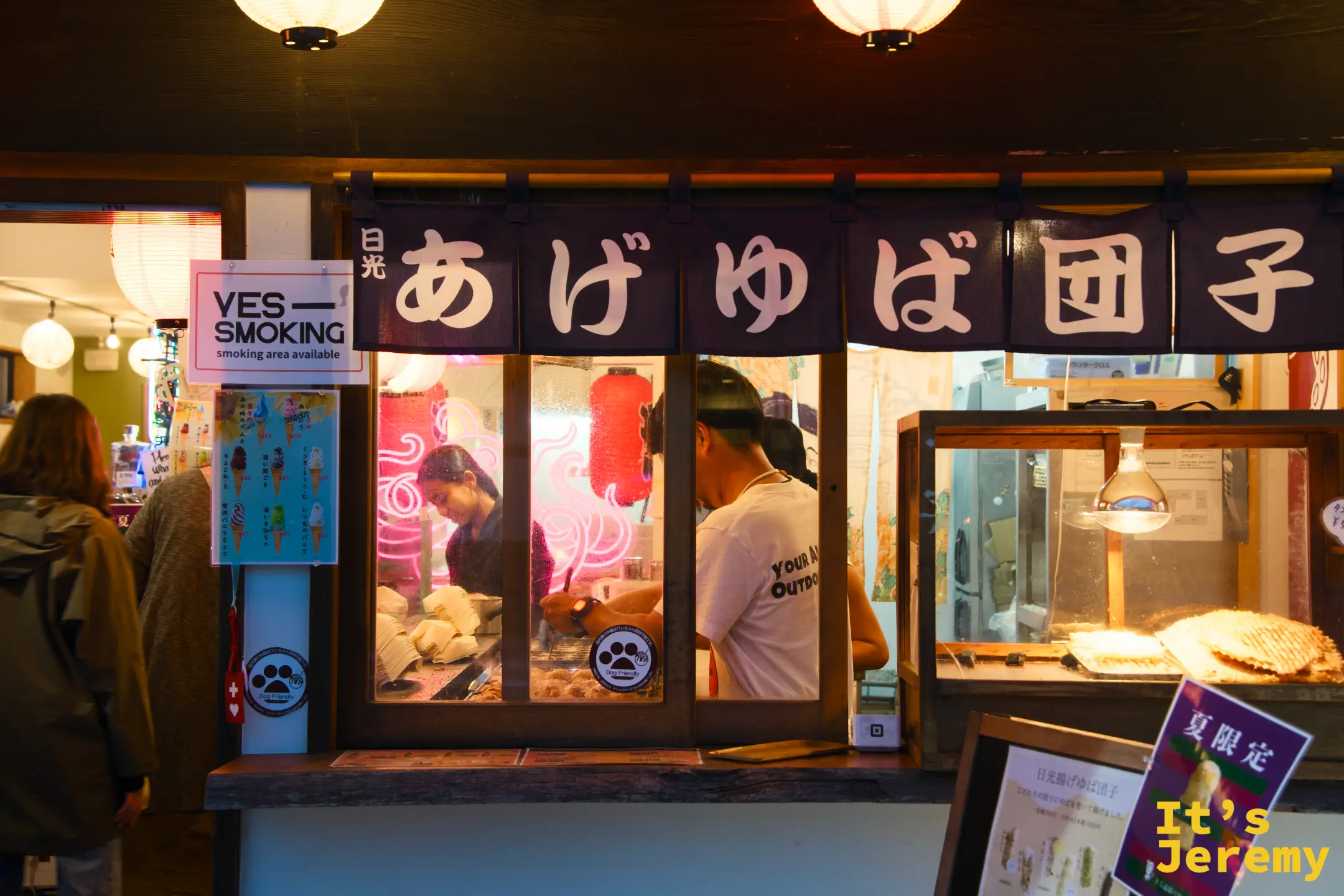 入夜的日光商店街，「あげゆば団子」攤位燈牌亮著，店員在攤前現做，遊客站在櫥窗前挑選