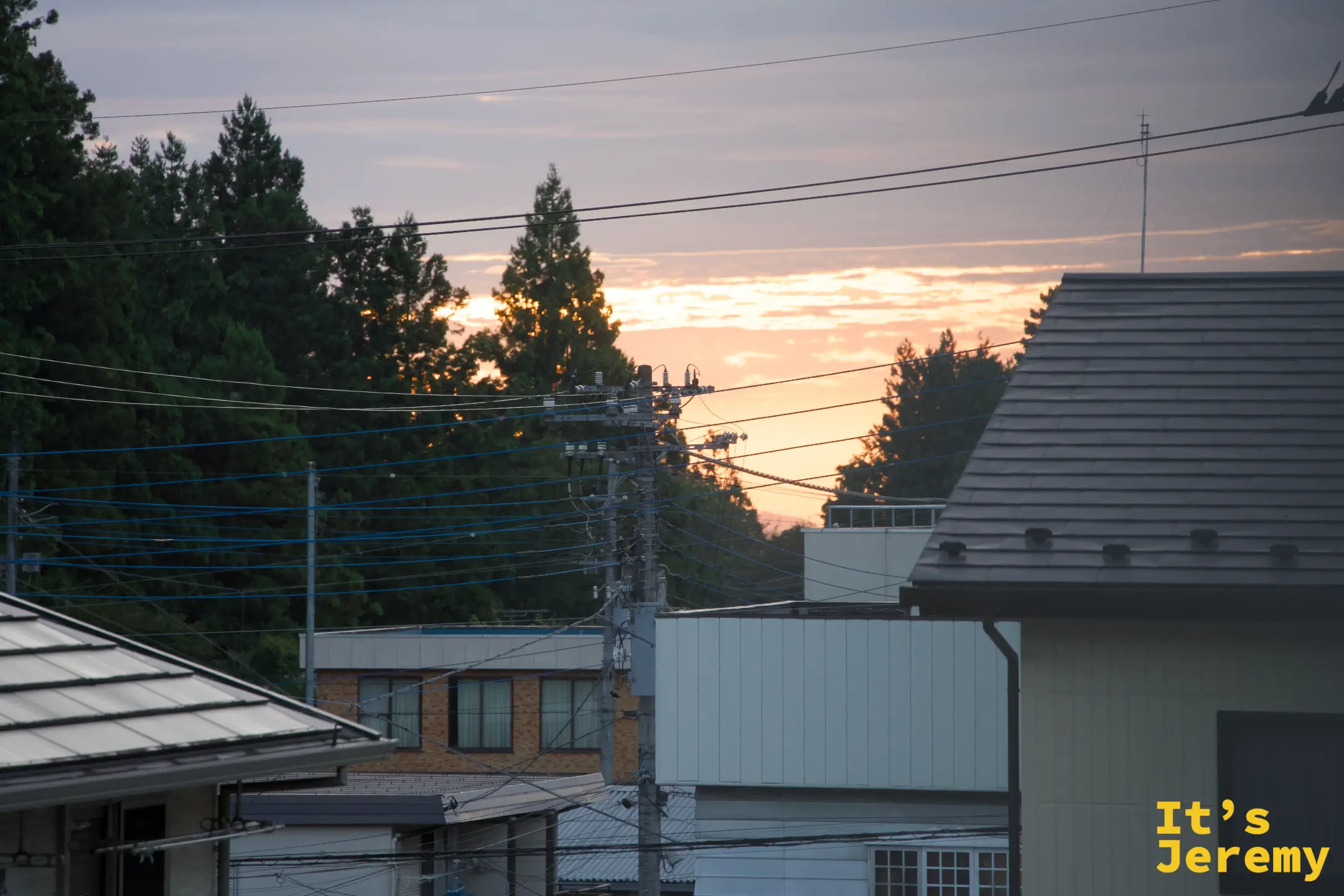 日光山城的傍晚，民宅屋頂與交錯的電線之間，夕陽把雲染成橘紅，杉木與遠山剪影連成一片