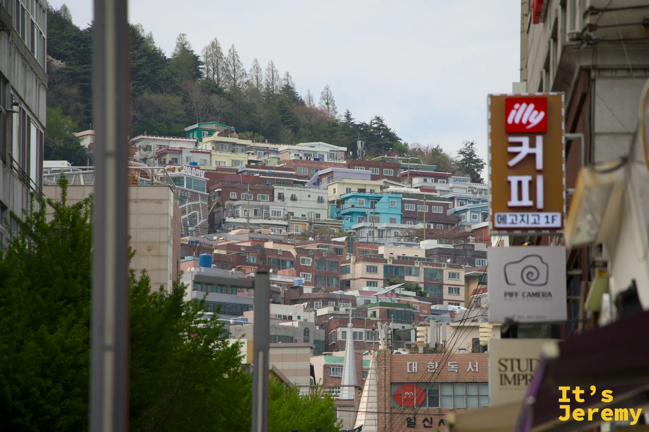 從街道往上望，山坡上密密疊疊的釜山住宅，前景是 illy 咖啡招牌和「커피」大字