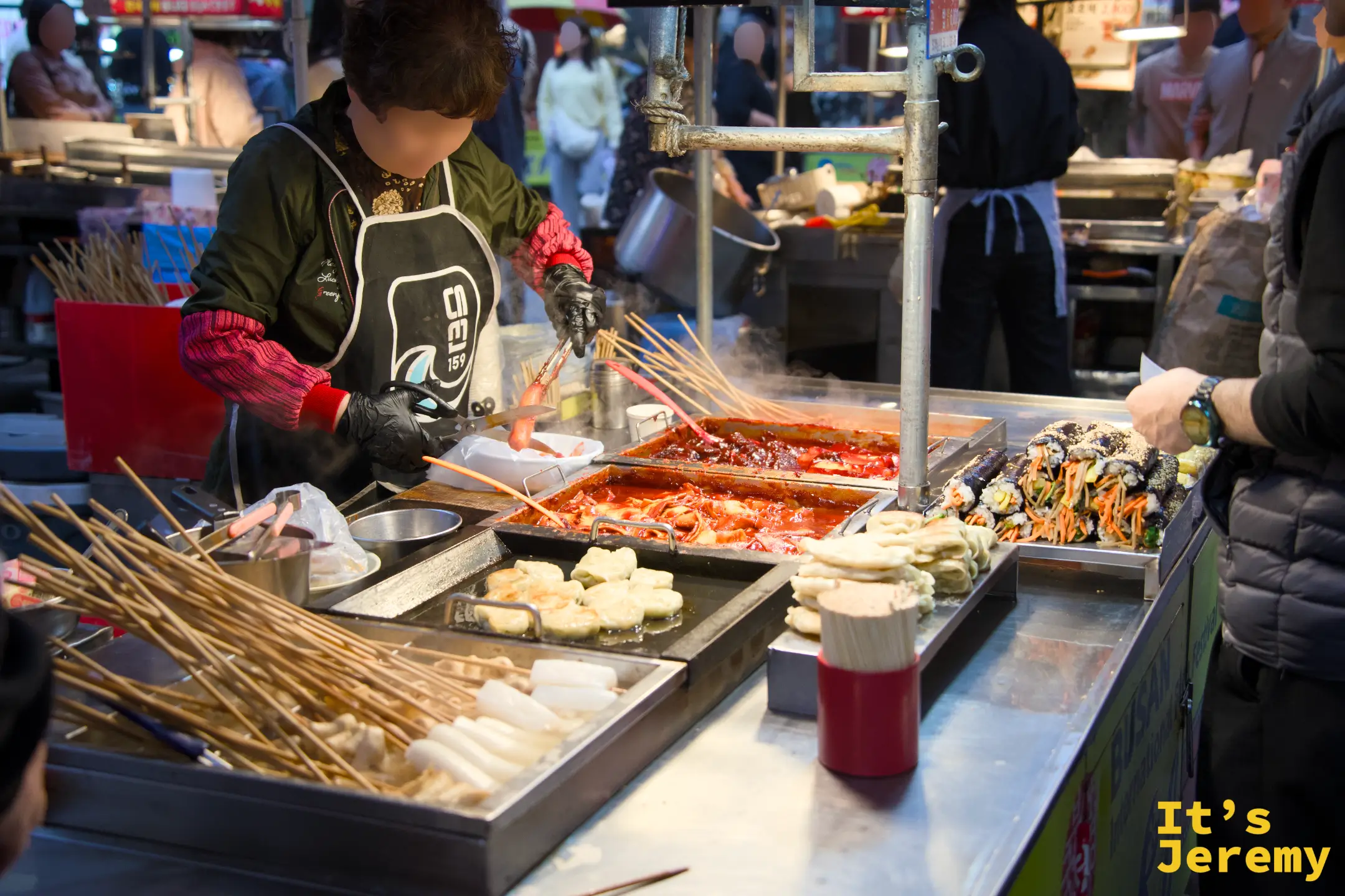 國際市場街食攤女老闆翻炒鐵板上的年糕、煎餃與海帶飯卷，熱氣四散