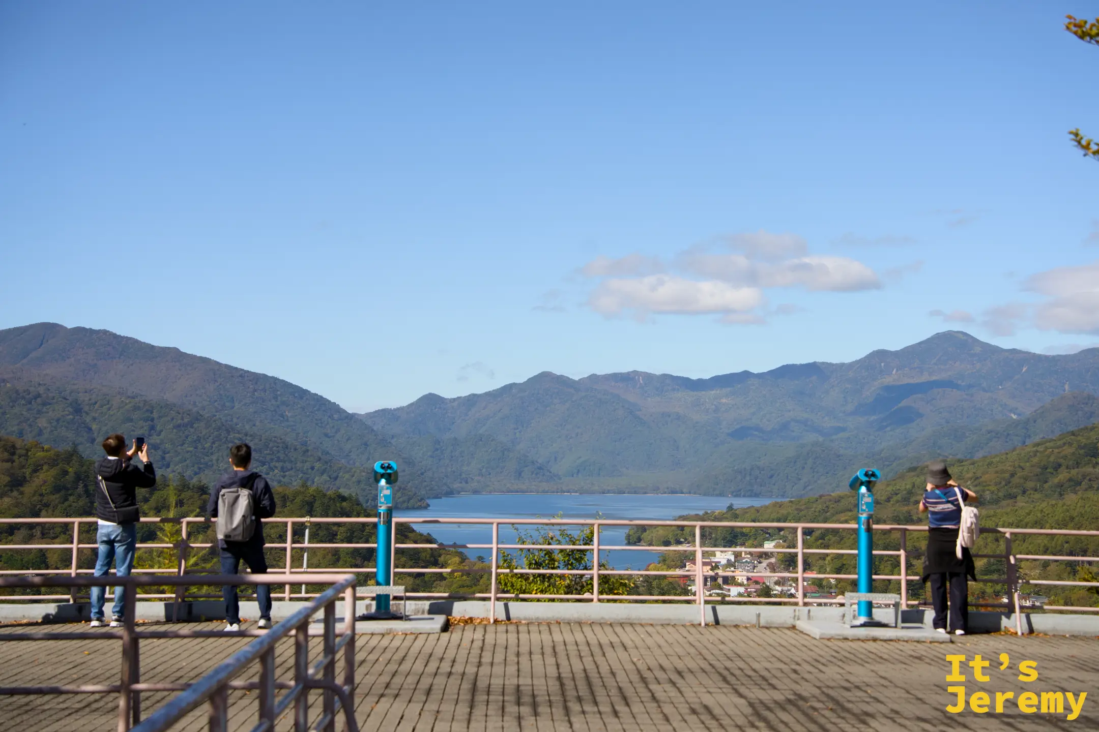 展望台觀景欄杆，遊客持手機記錄眼前的中禪寺湖全景，投幣望遠鏡立在兩側