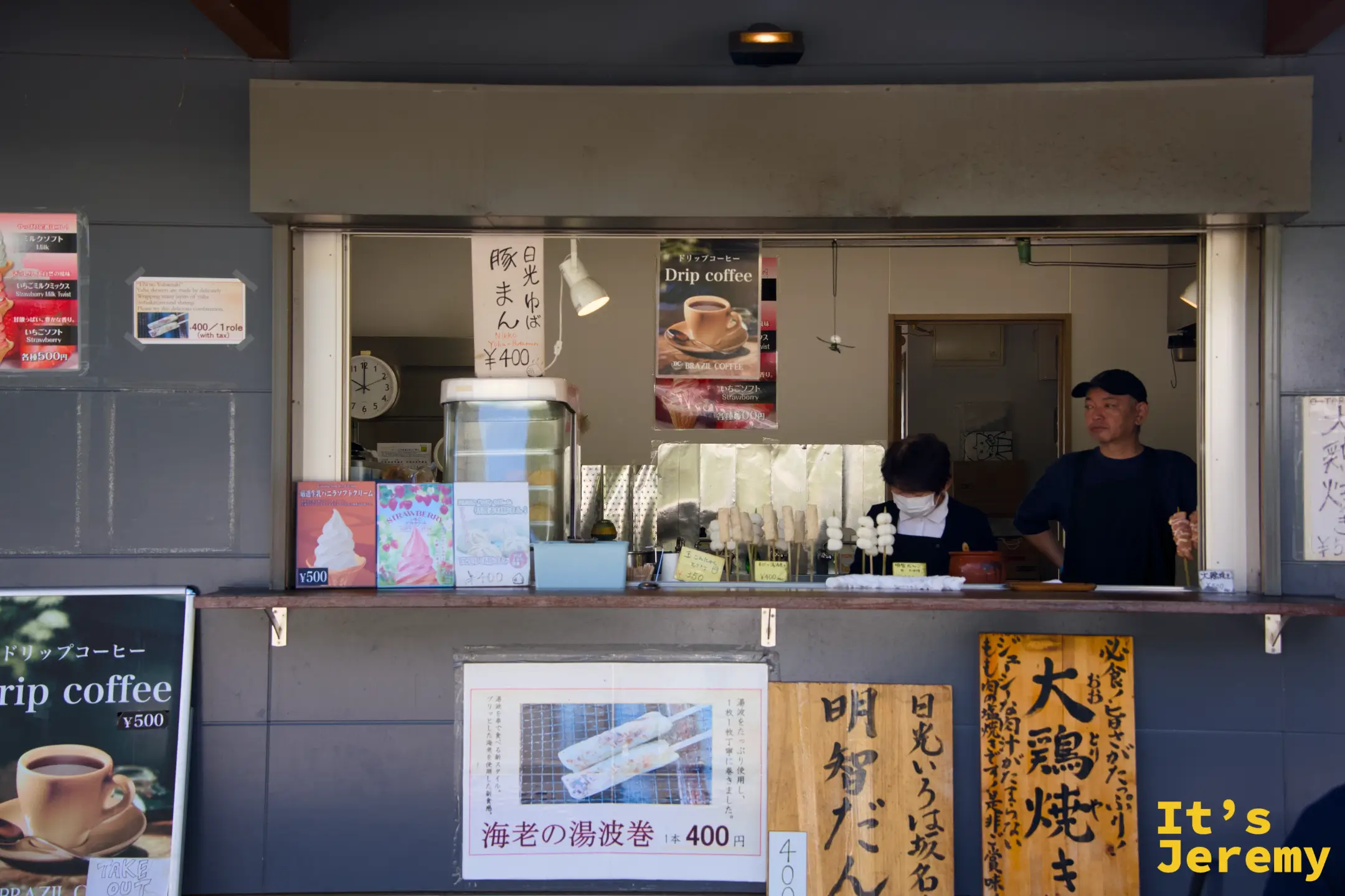 明智平賣店吧台，菜單上可見明智だんご、えびの湯波巻き、大鶏焼き等品項，兩名店員在內備料