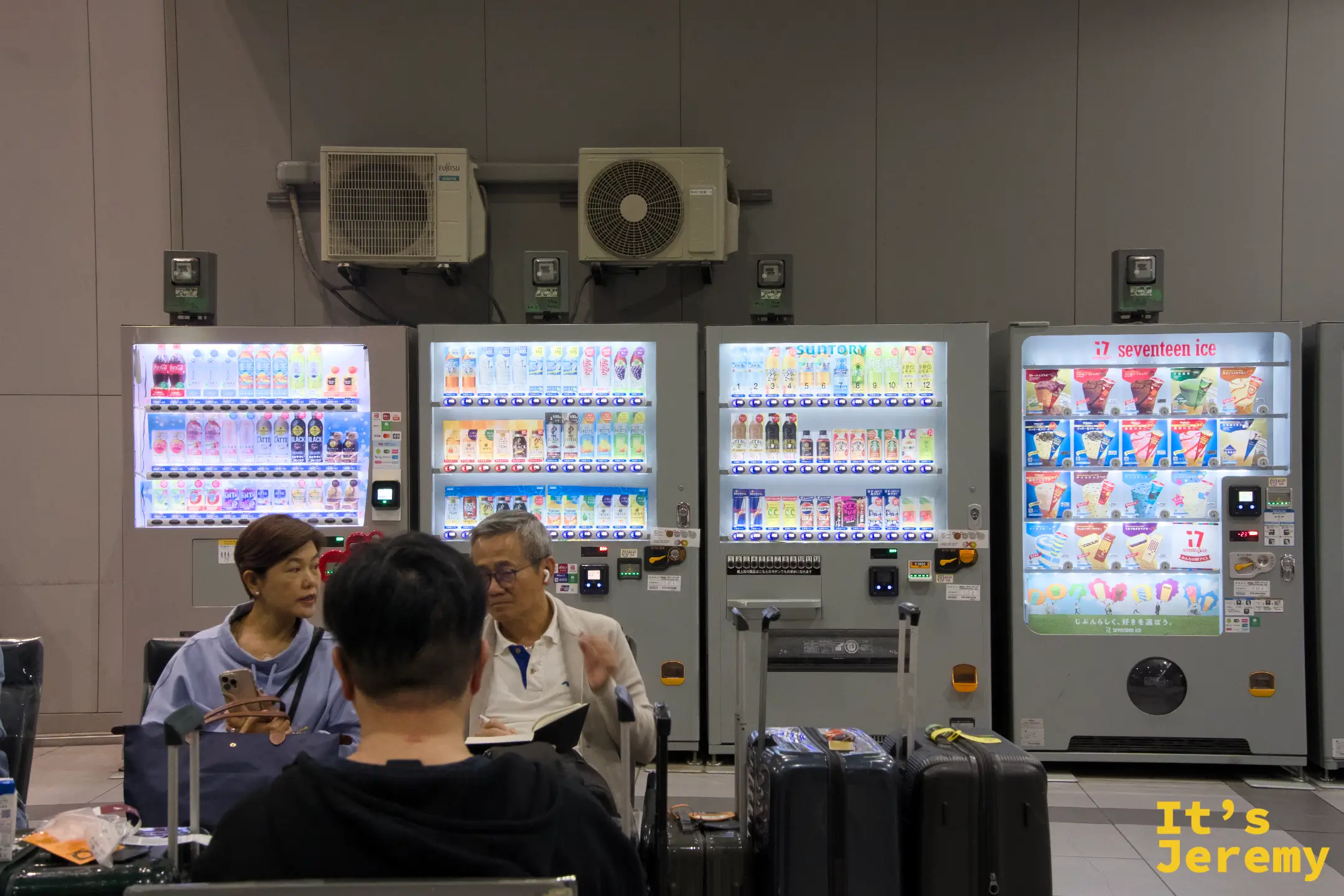 關西空港候車廳內，旅客坐在自動販賣機前閒聊，行李放在腳邊