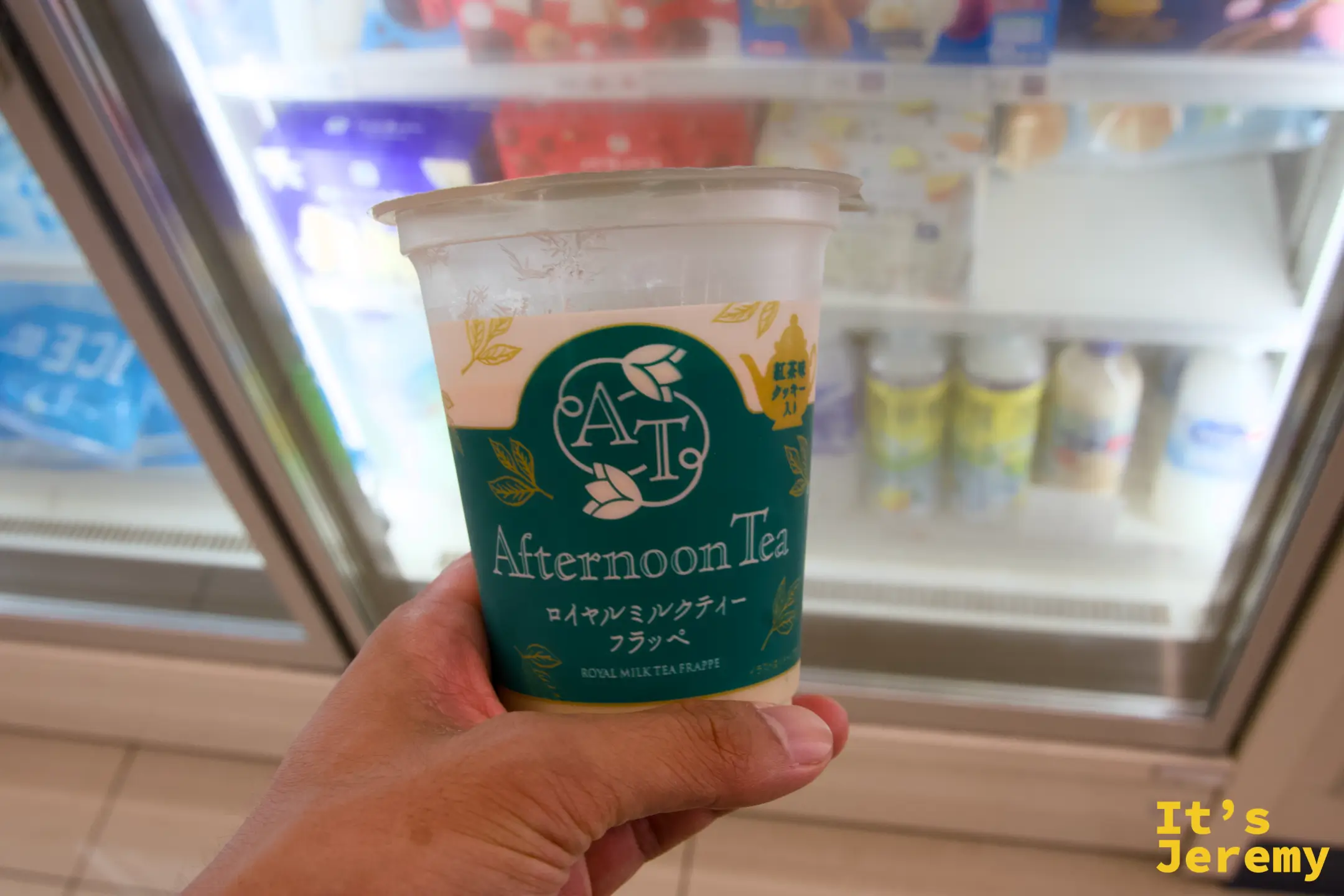 在全家冰箱前手持 Afternoon Tea 聯名「ロイヤルミルクティーフラッペ」，背景是各式冰品