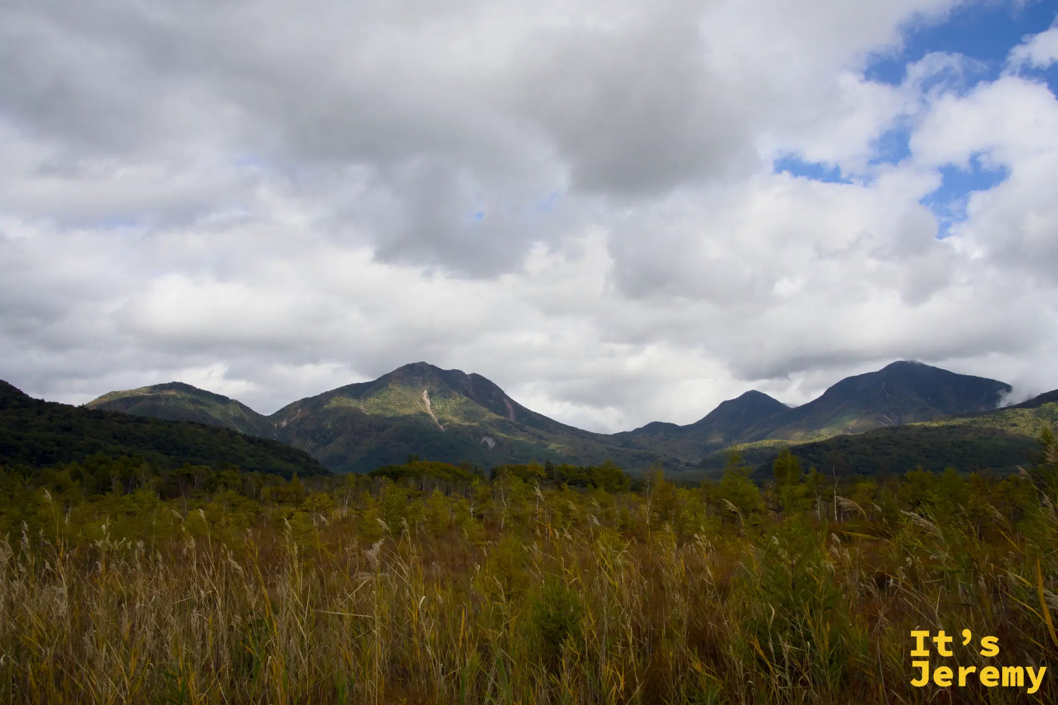 戰場平原廣角景色，金棕草原連接到遠處山脈，天空雲層厚重但仍有藍色間隙