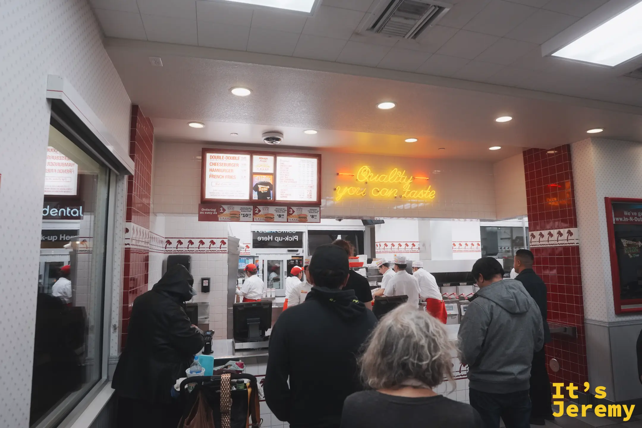 In-N-Out 點餐櫃檯，牆上菜單看板寫著 Double-Double、Cheeseburger、Hamburger、French Fries，店員穿白衣紅圍裙在煎台後方作業